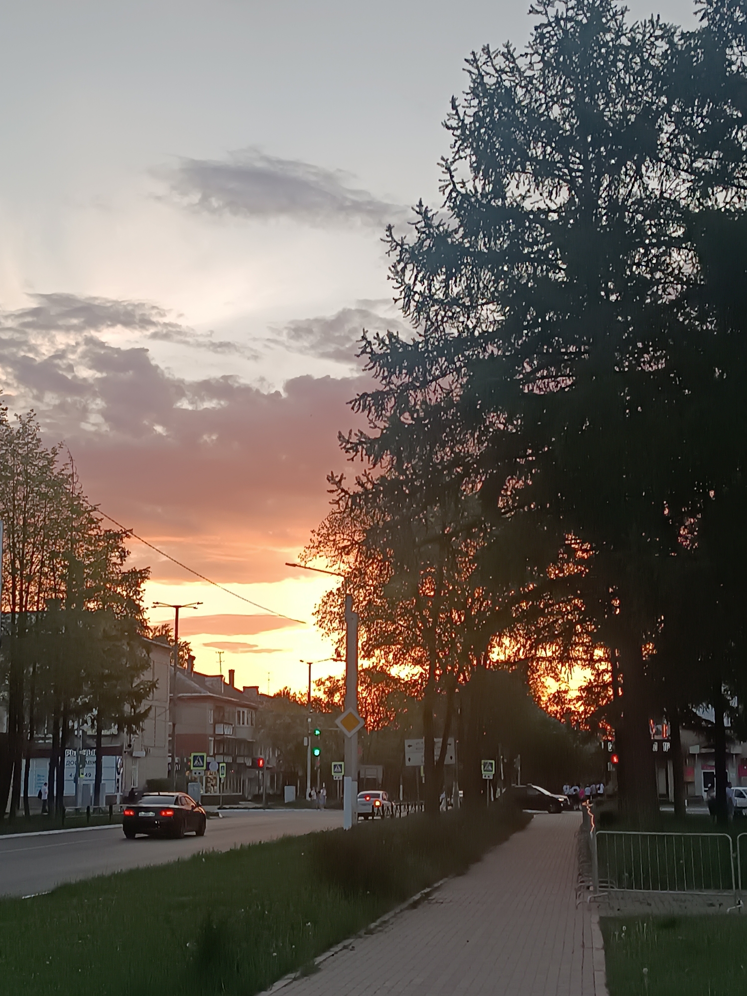 The image captures a sunset view along a street in what appears to be Chaikovsky, Russia. The sky displays a mix of colors, with a predominant orange and pink hue near the horizon, gradually fading into lighter tones higher up. Sparse clouds are scattered across the sky.
A road runs parallel to the viewer, with a car traveling along it. Buildings are visible on the far side of the road, interspersed with trees. Traffic lights are present, some showing red and others green, indicating active road intersections. Street signs are visible but illegible.
A sidewalk extends from the bottom right corner, made of rectangular paving stones. The foreground features patches of grass. A fence-like railing is situated next to the sidewalk.
Silhouetted trees dominate the right side of the image, their dense foliage obscuring details behind them. The overall lighting is subdued due to the sunset, creating shadows and emphasizing the silhouettes of objects and structures.