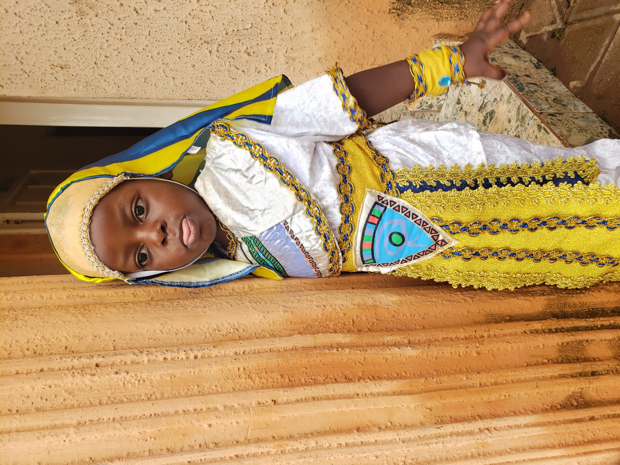 A young Black child is standing next to a textured orange wall, leaning slightly into it. The child is wearing a white and gold elaborate costume with blue accents. The costume includes a headpiece, a shimmering white tunic with golden trim and intricate patterns around the neckline and sleeves, a wide gold belt with blue trim and decorative tassels, and a matching gold sash adorned with colorful geometric shapes and a central blue circle. The child's left arm is extended, wearing a stack of yellow and blue beaded bracelets, with one blue bead prominently visible. The child is sticking their tongue out towards the camera. The background consists of the orange wall, a partial view of a doorway, and a tiled floor transitioning to dirt. The lighting is natural and suggests an outdoor setting in Sabon Tasha, Nigeria.