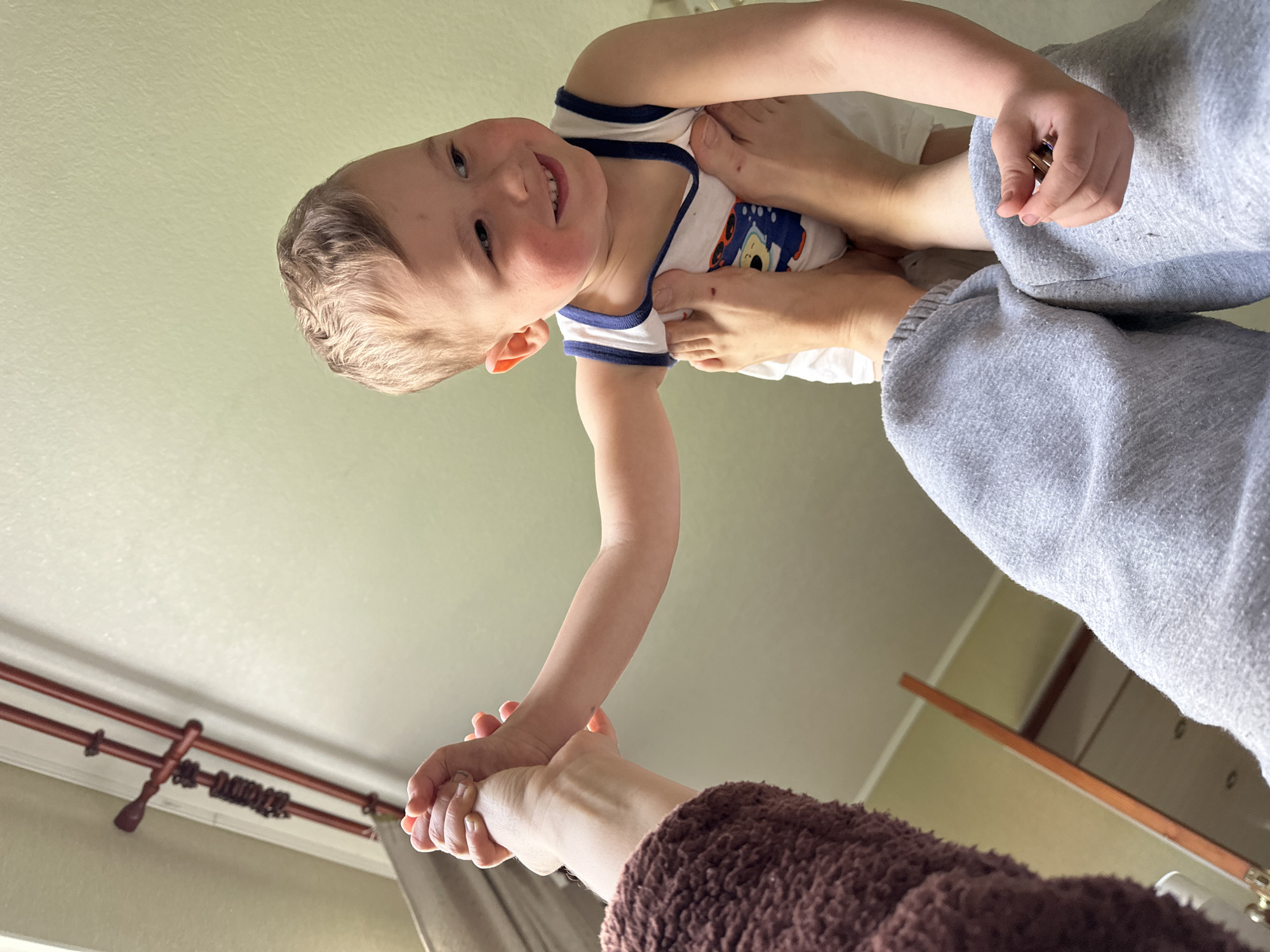 A young child wearing a white tank top with blue trim and a cartoon graphic is held upside down. An adult holds the child by the hand and feet. The adult wears grey sweatpants and a brown, fuzzy sweater. The background includes a light green ceiling, a dark brown curtain rod, and neutral-colored walls. The location is not apparent in the image.