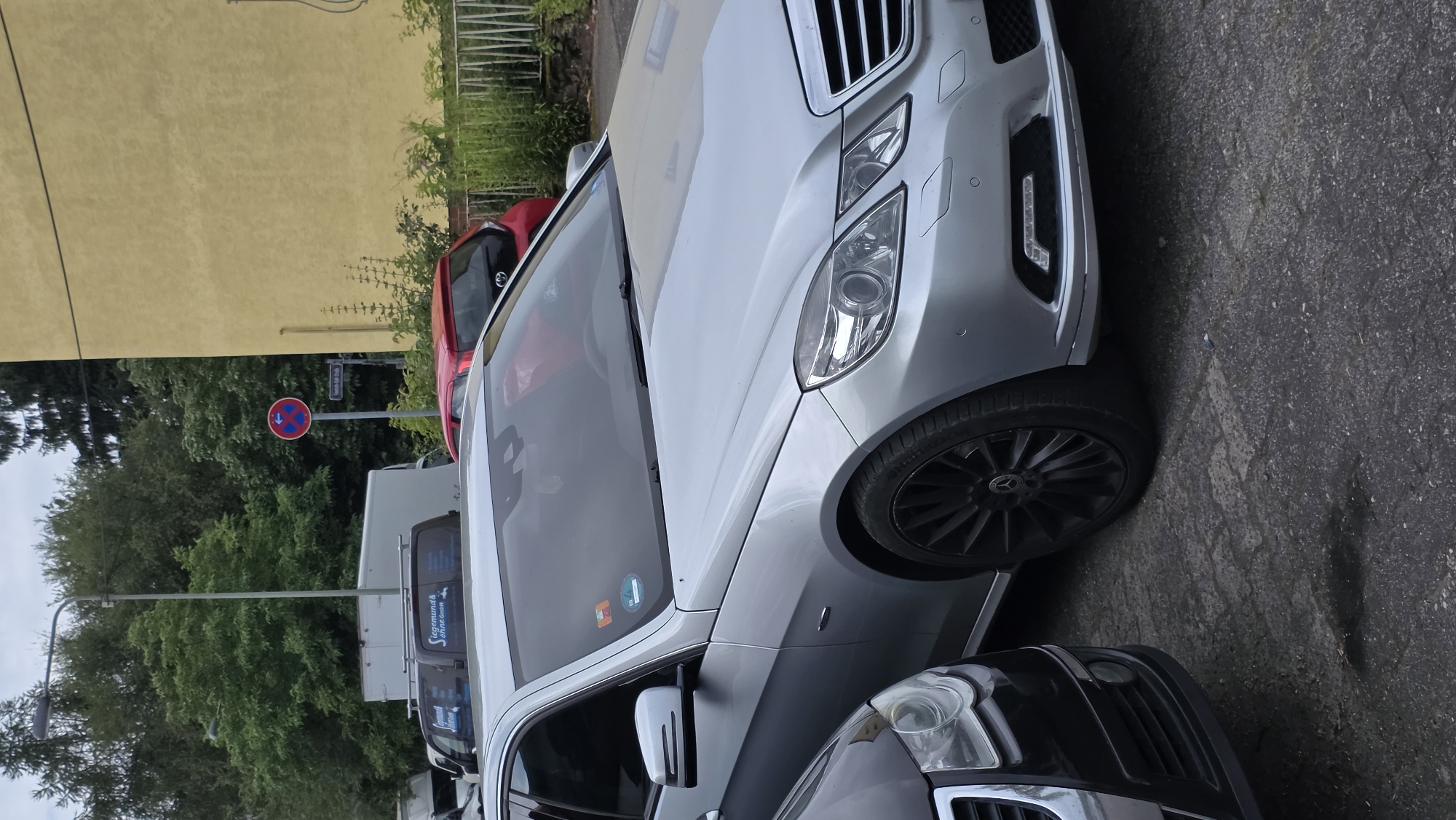 The image captures a close-up view of the front section of a silver Mercedes-Benz E-Class sedan parked on a paved surface. The car is positioned in the foreground, with its front bumper, headlights, grille, and front passenger side wheel clearly visible. The wheel is a distinctive black multi-spoke design. To the left of the Mercedes, the front end of a black car, possibly a Volkswagen, is partially visible, showing its headlight and bumper.
In the background, several other vehicles are present, including a white delivery van with "Siegemund & Ohrt GmbH" branding and a red hatchback. Behind the vehicles, a tall, yellow building with a rough textured wall dominates the right side of the frame, and a dense line of green trees forms the backdrop on the left and center. A street sign is visible on a pole, and a circular traffic sign with a red border and a blue arrow on a white background, indicating a no-parking zone, is also present. The sky is overcast, suggesting a cloudy day. The scene appears to be an urban or suburban street.