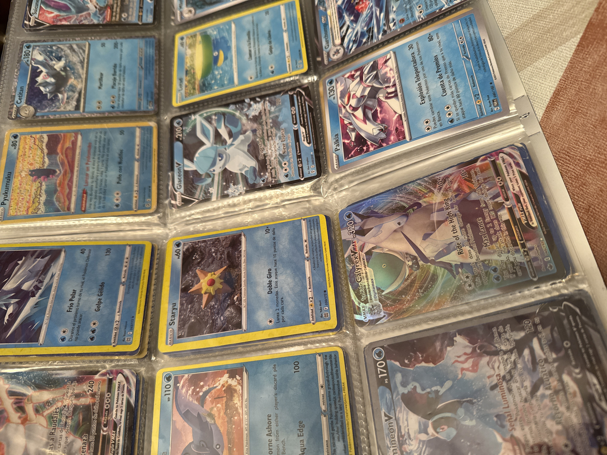 The image shows a binder filled with Pokémon cards. The cards are arranged in plastic sleeves. Several cards are visible, including Cetitan, Pyukumuku, Glaceon V, Staryu, Palkia, Ice Rider Calyrex VMAX, and Inteleon V. The cards are in Spanish. The background is a blurry, light brown surface. There is nothing in the image to indicate a location in Barcelona, Spain.