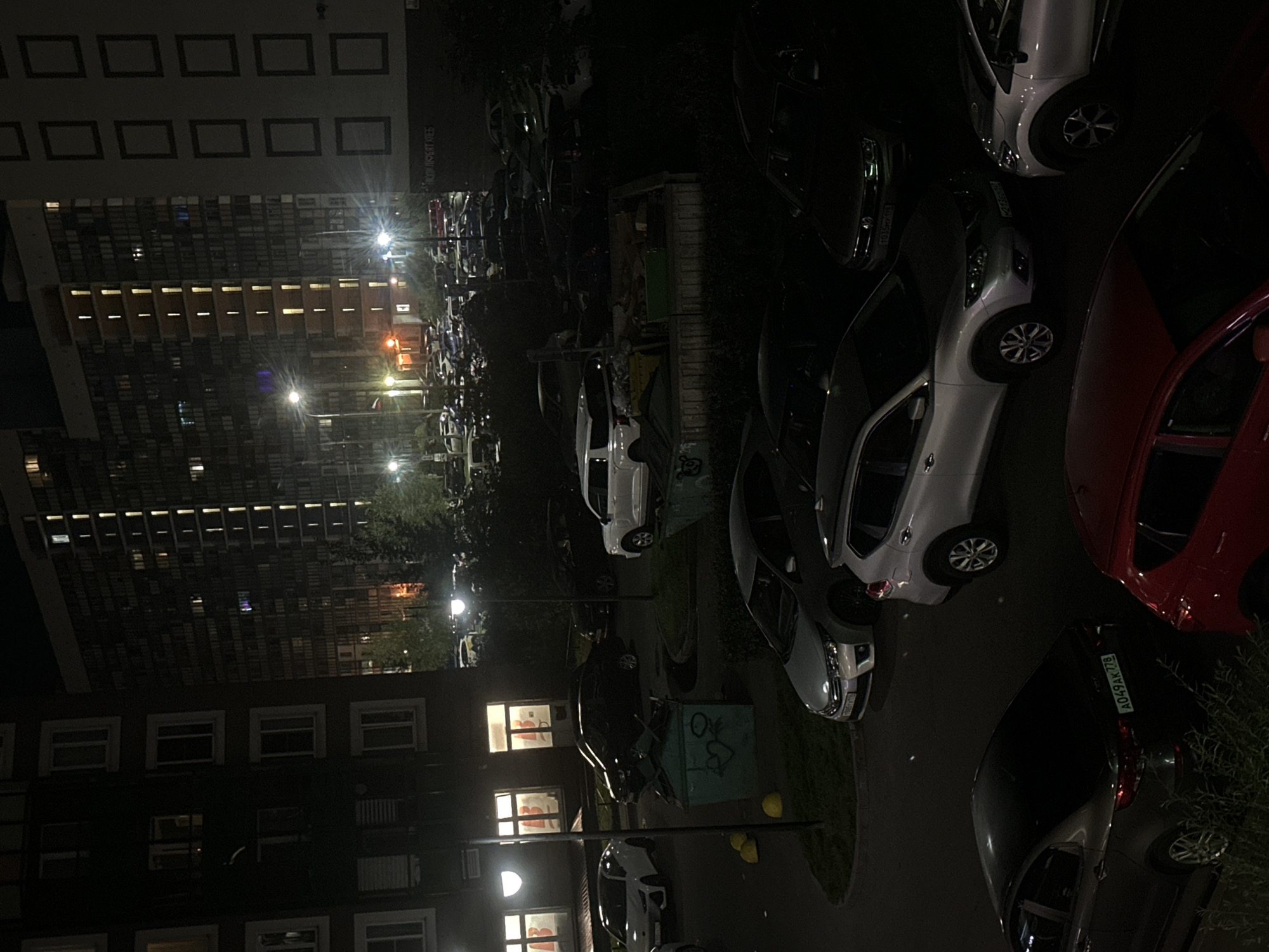 A nighttime, eye-level shot captures a residential scene in Sinyavino, Russia, focusing on apartment buildings and parked cars. Two tall, multi-story apartment buildings dominate the background, their windows illuminated with warm yellow light, suggesting occupancy. In the foreground and middle ground, numerous cars are parked on asphalt and grass areas. Vehicles include a white SUV, a silver sedan, a dark sedan, and a red hatchback, among others. Several large green dumpsters are visible, one with graffiti depicting a heart and an arrow. Streetlights cast pools of light, illuminating the parked vehicles and surrounding grassy areas. The environment appears to be a typical urban residential courtyard.