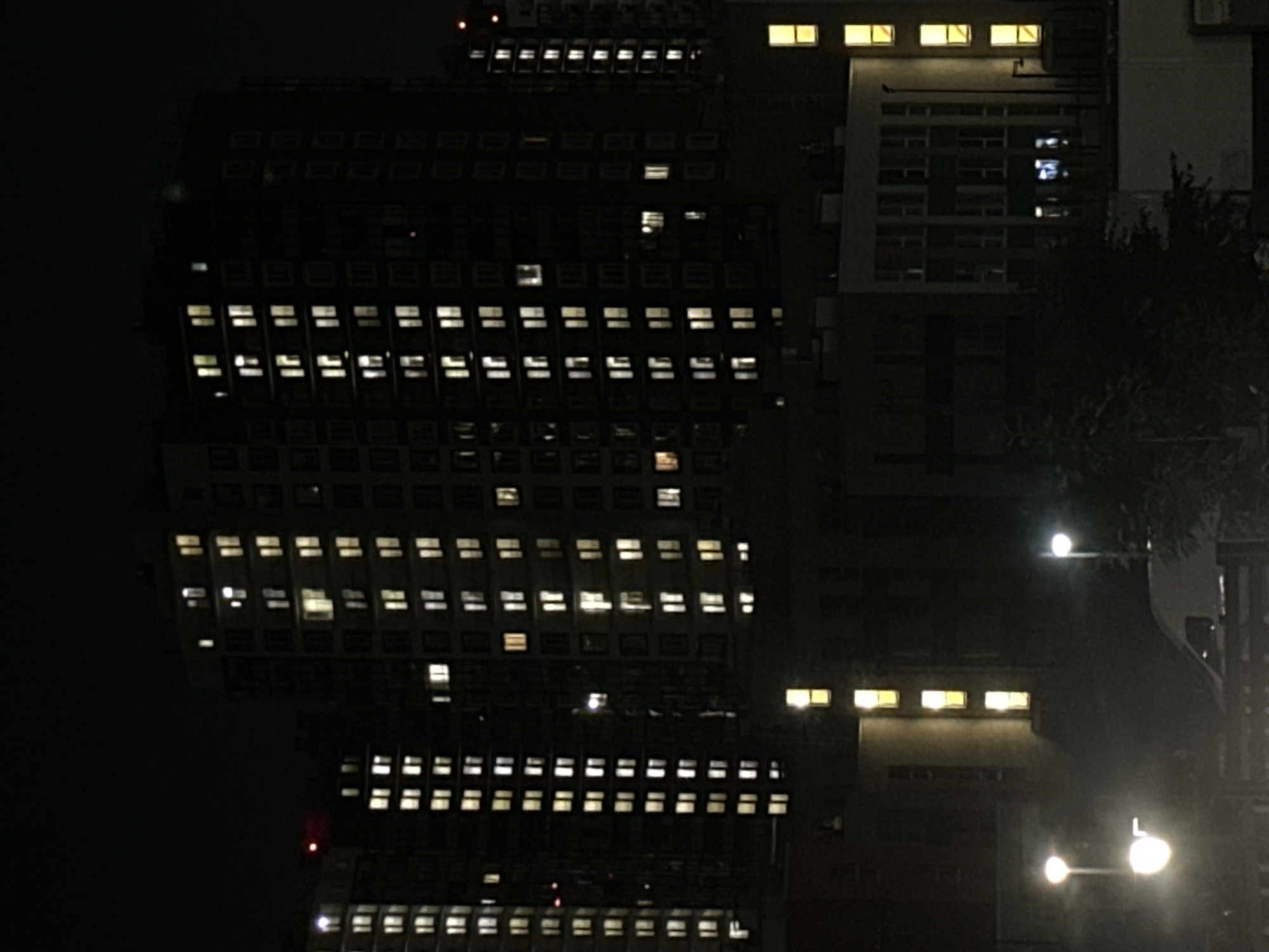 A night time aerial photograph captures a dense urban landscape dominated by apartment buildings. The structures are tall and rectangular, with numerous windows illuminated by artificial light, suggesting occupancy. The buildings are clustered together, with some appearing to be under construction or newer than others. In the foreground, streetlights cast a soft glow, and a portion of a white van is visible. Trees are scattered throughout the scene, adding a touch of nature to the concrete jungle. The overall impression is one of a bustling city at night, with life continuing behind the lit windows. The photograph provides a snapshot of urban living in Sinyavino, Russia.