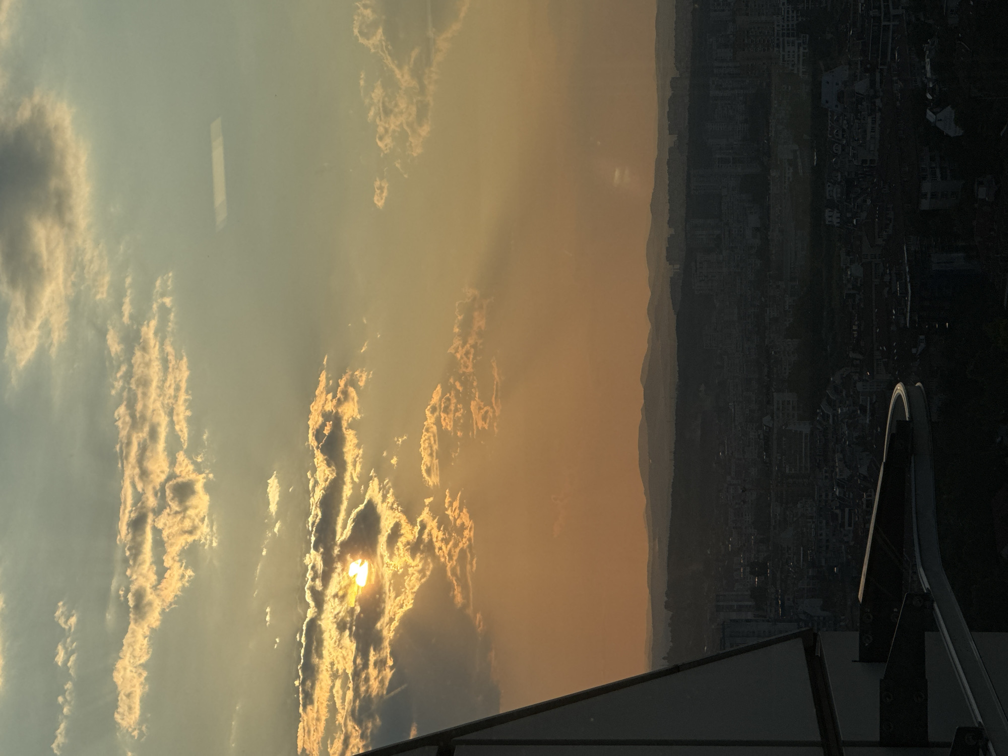 The image captures a sunset over Sofia, Bulgaria, viewed from a high vantage point, likely a building or observation deck. The sun is partially obscured by clouds, casting a warm, golden light across the sky. Rays of sunlight pierce through the clouds, creating a dramatic effect. Below, the city of Sofia sprawls across the landscape, a dense collection of buildings and structures, silhouetted against the colorful sky. In the foreground, a modern architectural element, possibly part of a building's facade or a viewing platform, is visible on the left side, with its geometric lines and reflective surfaces contrasting with the natural beauty of the sunset and the urban panorama. The overall impression is one of a serene and expansive urban sunset.