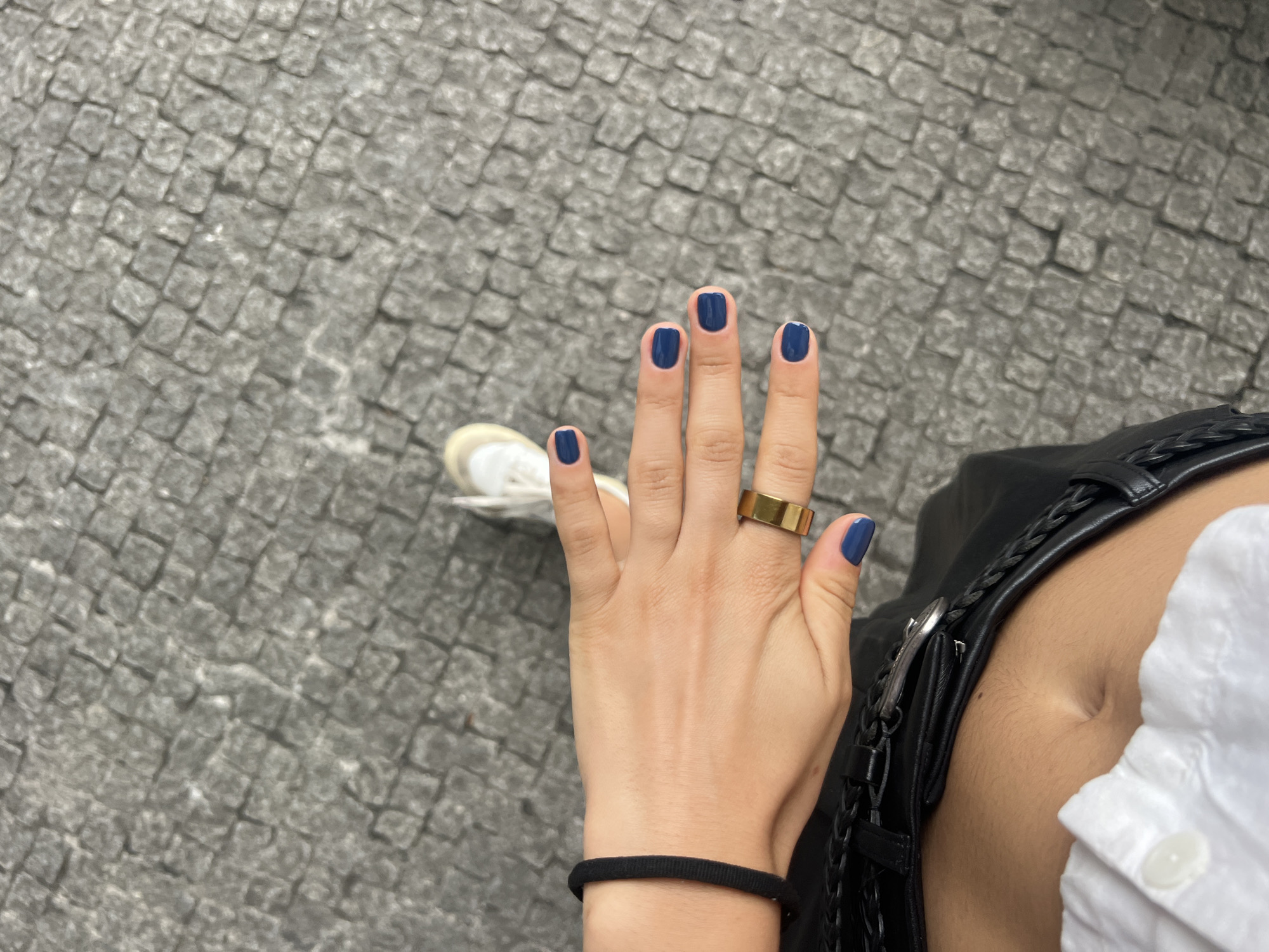 A first-person perspective shot shows a hand with dark blue nail polish and a gold ring. The hand is in the foreground, extended towards the viewer, with cobblestones visible in the background. Part of a white shirt, black leather pants, and a white sneaker are also visible. The setting appears to be a street in Porto, Portugal, characterized by the traditional cobblestone pavement.
