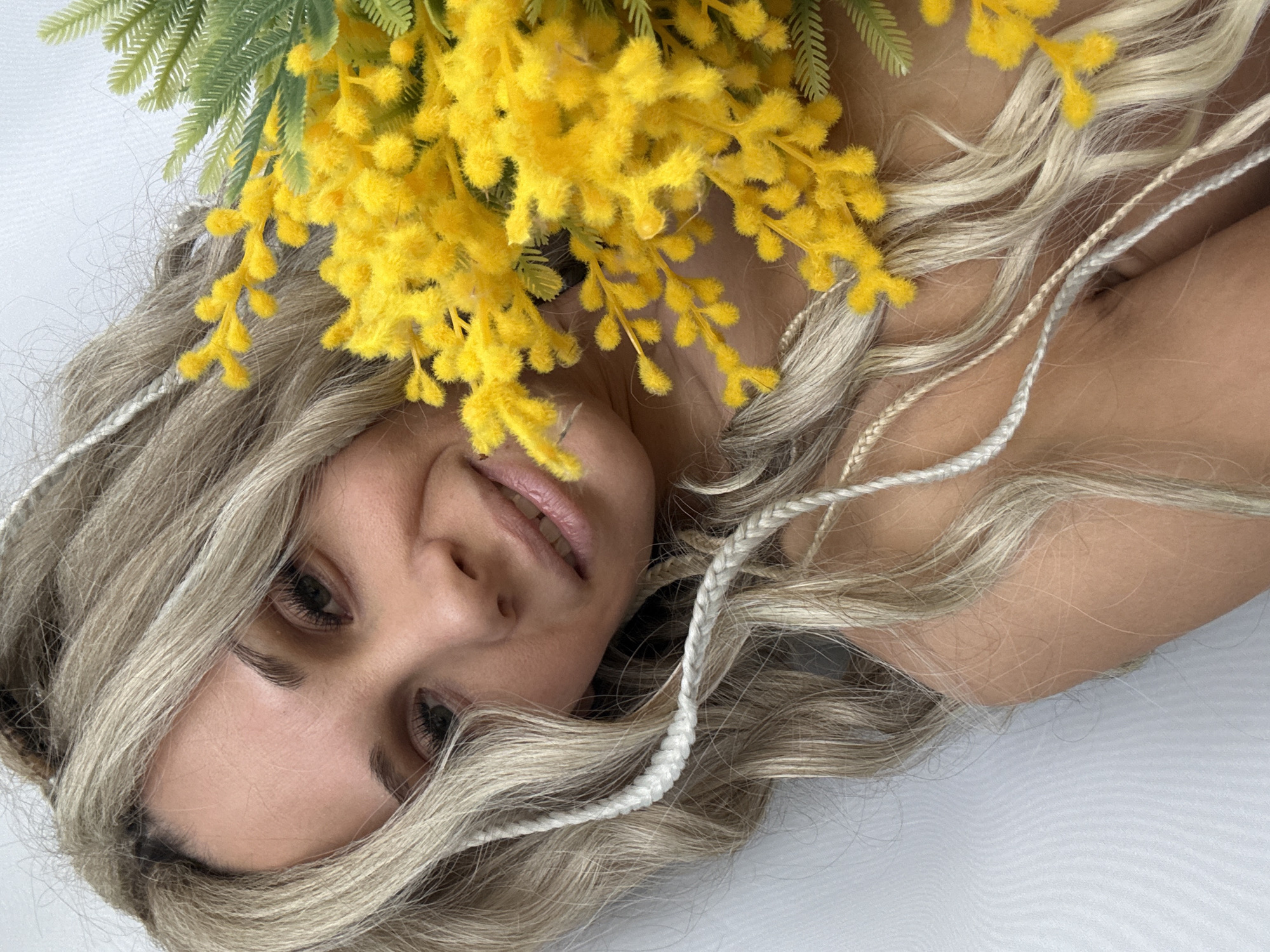A medium shot depicts a blonde woman with gray-blue eyes partially obscured by yellow Mimosa flowers. The flowers, with green stems and foliage, rest on her head. Her wavy, shoulder-length blonde hair is intertwined with two white braids. The woman's head is tilted, and she gazes upwards towards the camera with a slight smile. The background is a smooth white surface. The scene is located in the city of Unknown, Unknown.
