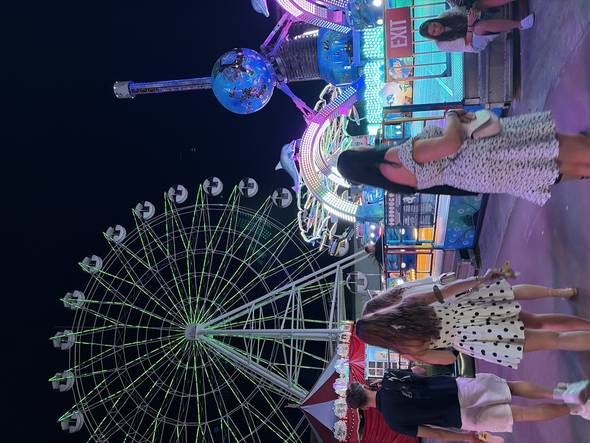 A group of people are walking through an amusement park at night in Nesebar, Bulgaria. A large Ferris wheel with green lights dominates the background, with a smaller ride featuring dolphins and colorful lights in the middle ground. The foreground shows a man in a black shirt and white shorts, followed by a woman in a white polka dot dress and another woman in a floral dress. An "EXIT" sign is visible on a booth to the right, and a girl in a white shirt and denim shorts sits on steps in the far right. The ground is paved, and the overall atmosphere suggests a lively evening at an entertainment venue.