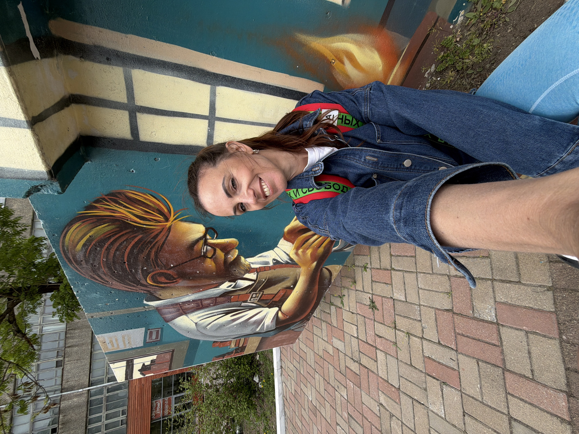 A woman in a denim jacket is smiling into the camera, holding it at arm's length. She is positioned to the right of a mural of a man with glasses, who is dressed in a white collared shirt, and suspenders. The mural is painted on a wall. The mural includes a depiction of a building behind the man. The background beyond the wall features a multi-story building with numerous windows and some greenery. The street is brick. The woman is likely standing in Birobidzhan, Russia, given the lettering on the mural.