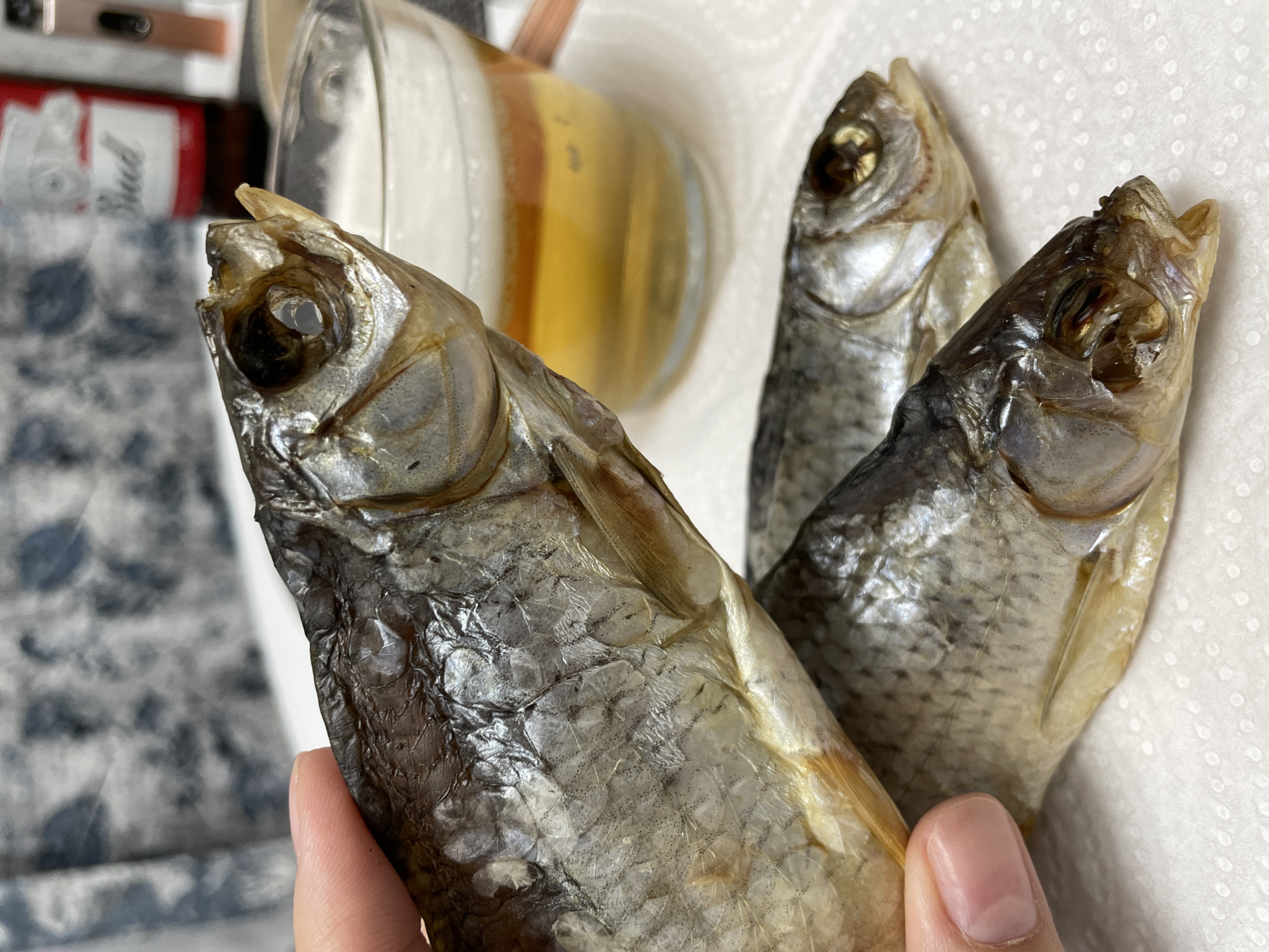 A close-up shows a hand holding three dried fish. The fish are small, with silvery scales and empty eye sockets. A glass of amber liquid is partially visible in the background. The background also includes a can with red and white labeling. The surface beneath the fish appears to be white paper towel. There is no verifiable information available to confirm this photo was taken in Izhevsk, Russia.