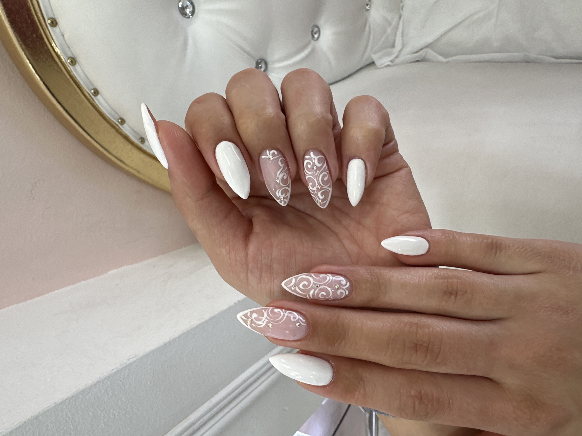 A woman's hands are displayed, showcasing a fresh manicure with white and nude-toned nail polish. Some nails are solid white, while others have a nude base with white swirling patterns and small rhinestones. The nails are shaped into a pointed almond or stiletto style. The hands are positioned against a background featuring a white tufted surface, possibly a headboard or chair, with a gold-trimmed, oval-shaped object visible in the upper left corner. The background also includes a white countertop or wall.
The location is identified as Sofia, Bulgaria.