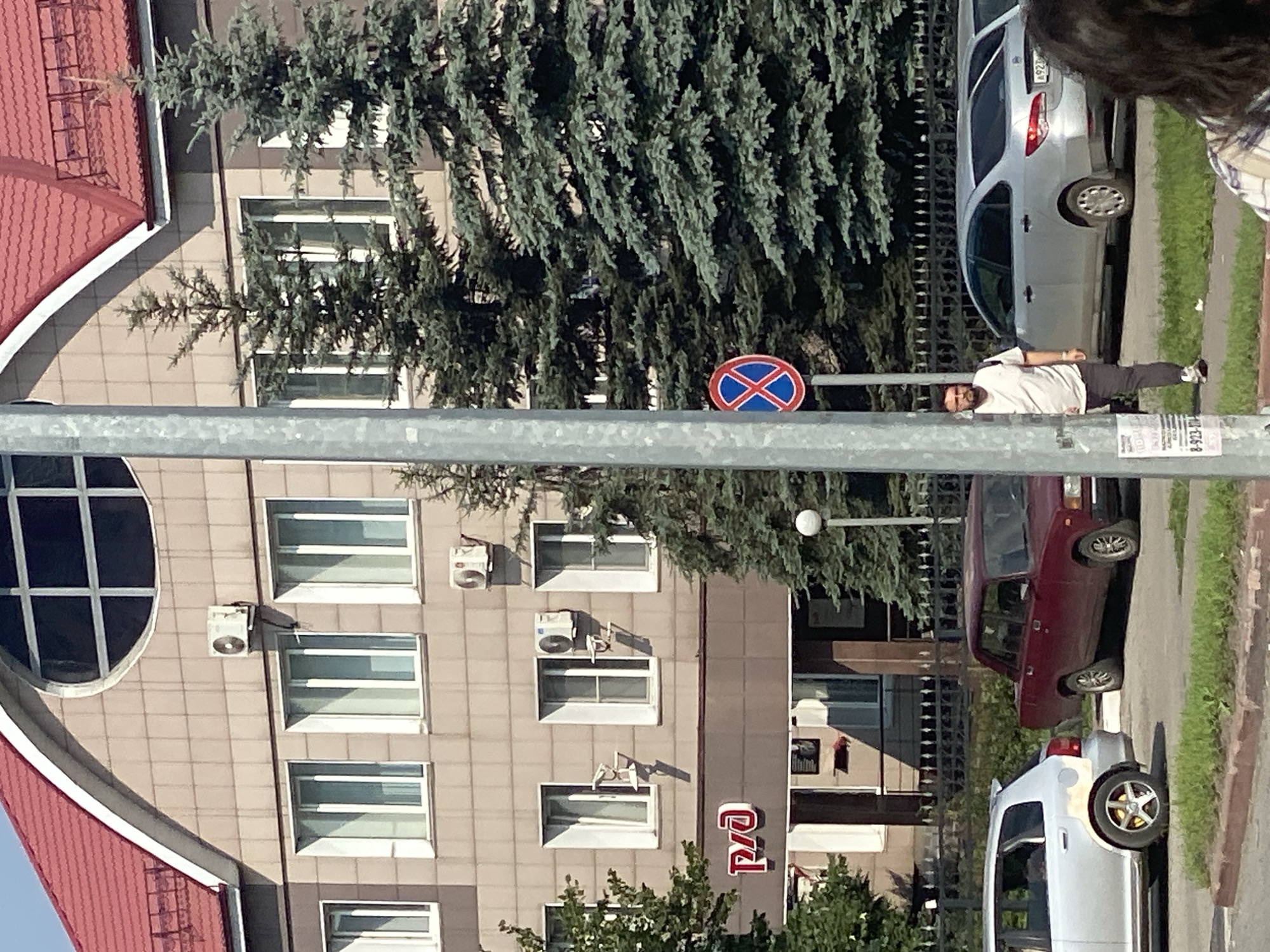 An exterior shot shows a man in a white t-shirt and dark pants walking past a building and trees. The building appears to be a multi-story office or administrative building with a red roof and beige tiled exterior. Air conditioning units are mounted on the exterior walls. Several windows are visible, and a sign with red Cyrillic lettering reading "РЖД" (RZD, Russian Railways) is mounted above an entrance. A round traffic sign with a red circle and a blue cross is mounted on a pole to the left of the man. Two cars are parked on the street in front of the building, a silver hatchback on the left and a maroon sedan on the right. A silver sedan is parked further down the street. The scene is set in Novokuznetsk, Russia.