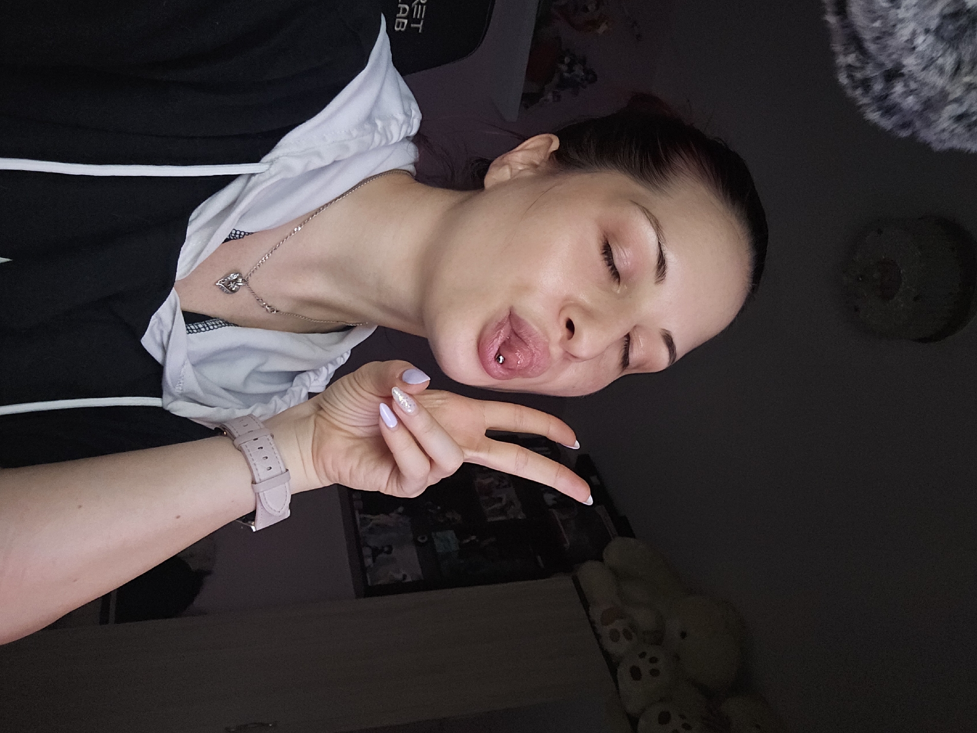 An image taken from a high angle shows a young woman in an indoor setting, likely a bedroom or living space, in dim lighting. She appears to be lying down or reclining, looking upwards at the camera. Her eyes are closed, and she is sticking out her tongue, which has a metallic piercing. Her dark hair is pulled back, revealing her face.
She is wearing a black hooded top with white trim and a white drawstring. Around her neck, a silver chain with a heart-shaped pendant is visible. Her left arm is raised, and her hand is making a "peace" sign. She wears a light pink watch on her wrist, and her fingernails are painted light purple with one nail featuring glitter.
In the upper left background, a black surface, possibly a chair or headrest, displays partially visible text that reads "RET". To the upper right of her head, a dark, textured, fluffy object (possibly a blanket or pillow) is seen. In the lower right corner, several light-colored plush toys with faces are partially visible. The scene has a relaxed and playful atmosphere, likely captured in the evening or at night due to the low light. The location is Burgas, Bulgaria.