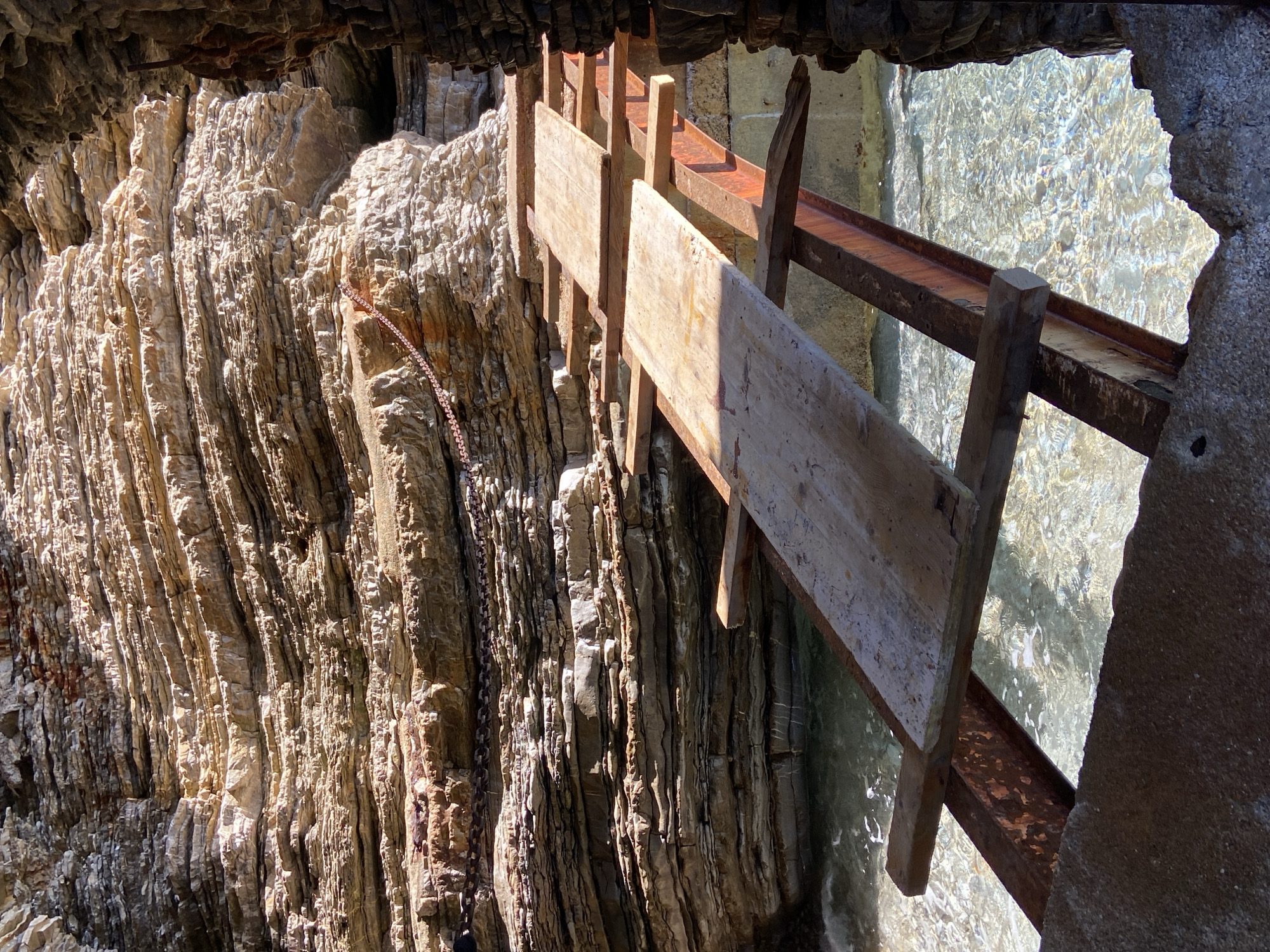 The image depicts a rock face with a weathered, light-colored, and layered texture. A chain hangs along the rock face. Overlapping the rock face is a weathered, wooden structure, possibly part of a water wheel or similar mechanism. A rusty metal beam is also visible. In the background, there is water and a part of a structure, potentially made of concrete or stone.
This scene is in Budva, Montenegro.