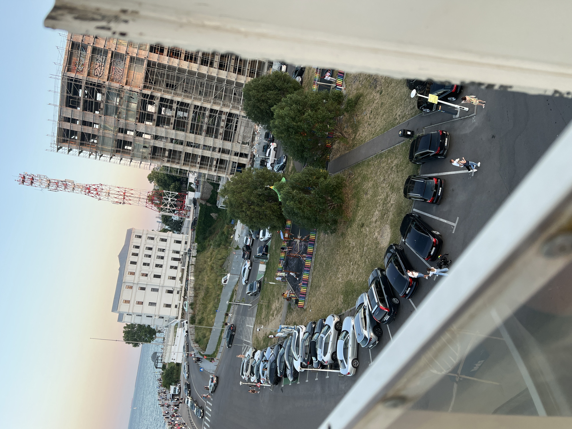 The image shows an aerial view of a street and parking lot in Constanța, Romania, near the Black Sea. Several cars are parked in a line, and a playground is visible to the right. In the background, there's a large white building and a construction site with a red and white radio tower. People are walking on the sidewalk and near the playground. The sky is clear with a light gradient of colors, suggesting it's either sunrise or sunset.
