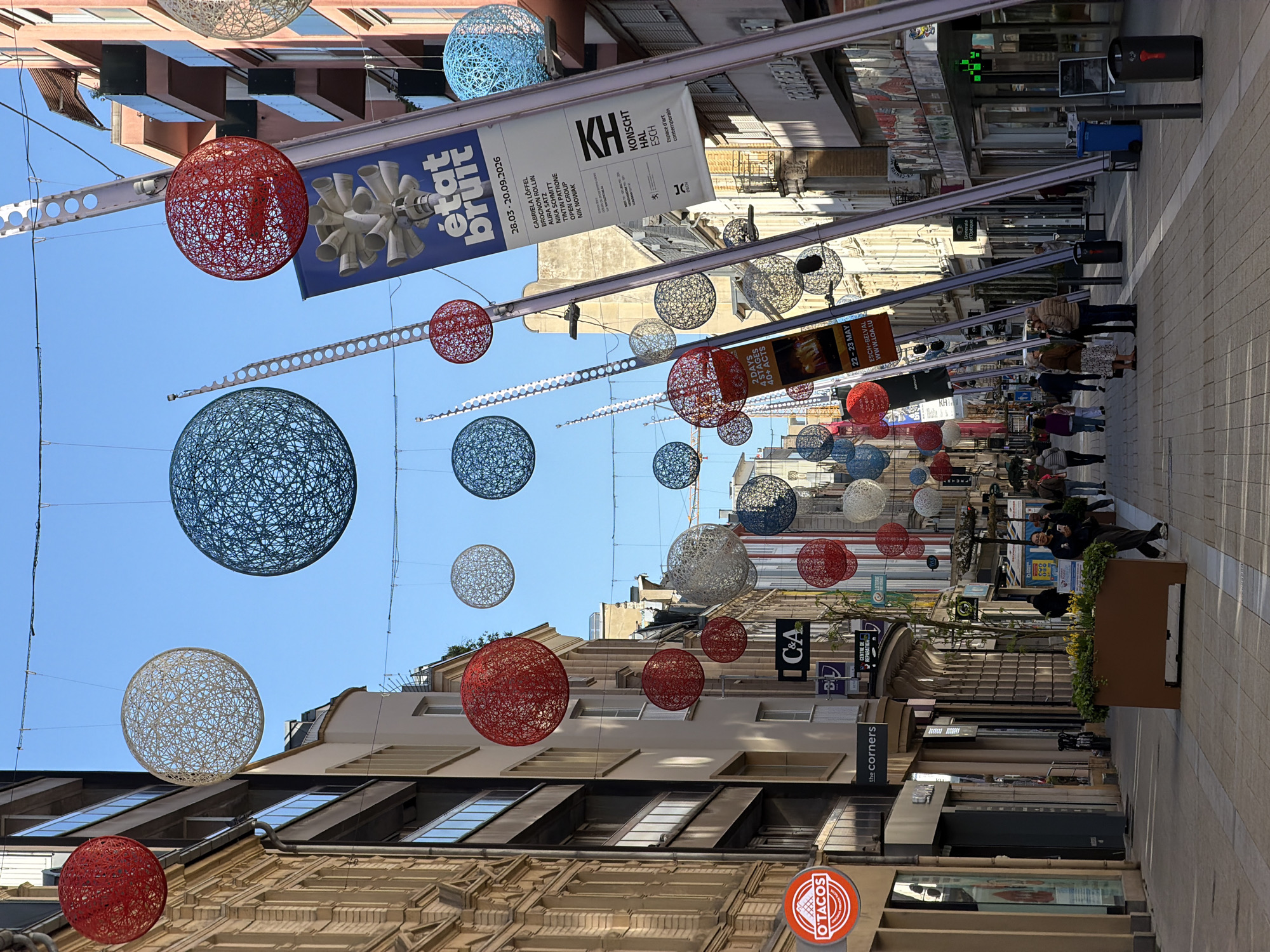 The image depicts a street scene in Esch-sur-Alzette, Luxembourg, characterized by decorations suspended above the pedestrian zone. These decorations consist of spherical, web-like structures in red, white, and blue. Banners are also suspended. Banners advertise the "état bruit" art exhibition at Konscht Hal Esch, running from March 28 to September 20, 2026, and an event called "2 Days 4 Stages 40+ Acts," from May 22-23 at Esch-Belval. People are walking along the street. Shops line both sides, including "O'TACOS" and "the corners." There's also an "IC&A" and "CENTRE DE REPARATION." A pharmacy with a green cross symbol is visible in the distance. Potted plants are placed along the street. The sky is clear and blue.
