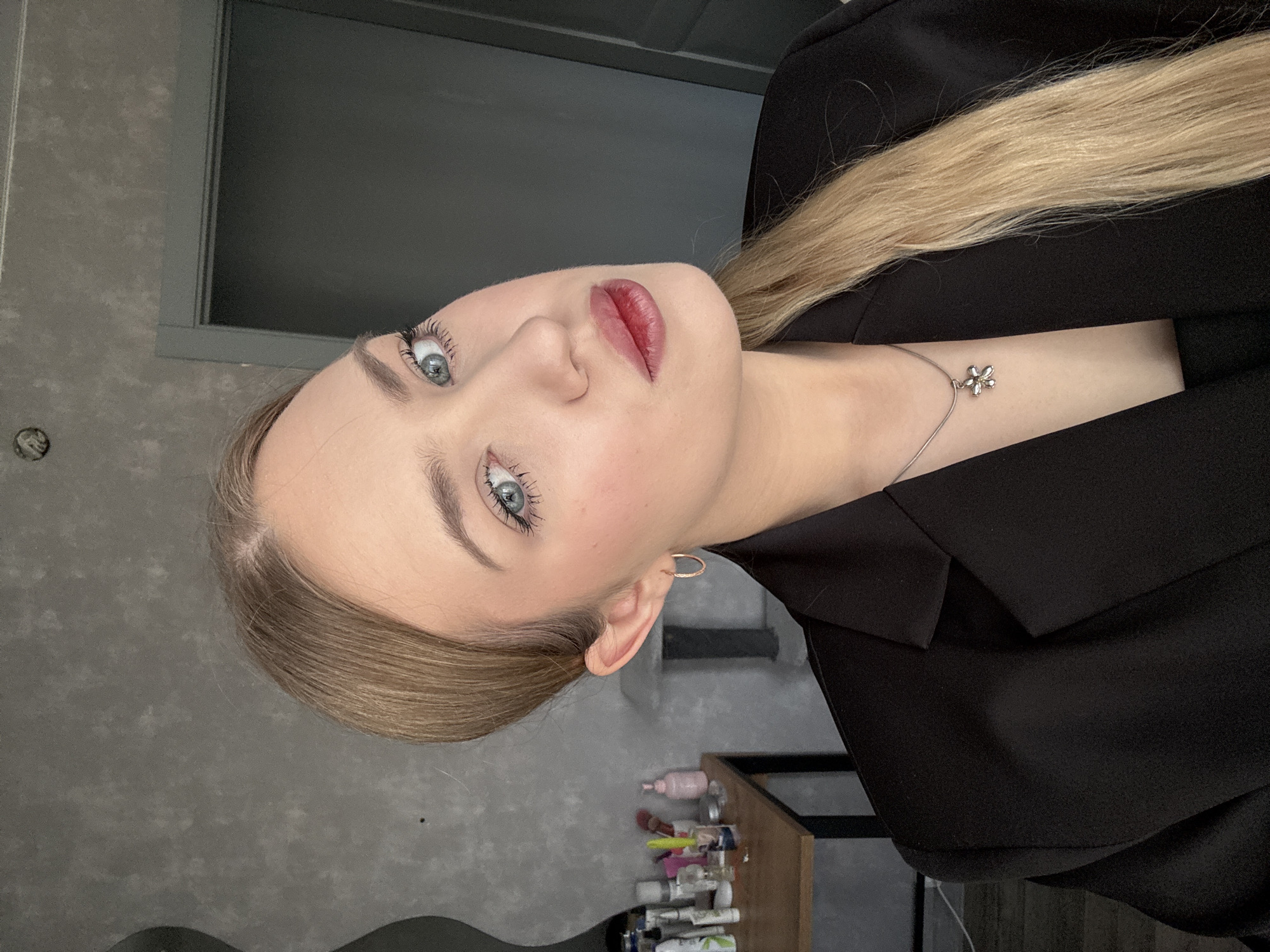 A person with light skin and blonde hair is seen in a close-up shot, angled towards the camera. The person is wearing a black blazer and a delicate silver necklace with a flower-shaped pendant. They have blue eyes and are wearing lipstick. An earring is visible on the right ear. The background appears to be a room with a gray wall and a window with a gray frame. In the lower right corner, a small wooden shelf holds an assortment of bottles and tubes. The image does not contain information confirming the location in the city of Chaikovsky, Russia.
