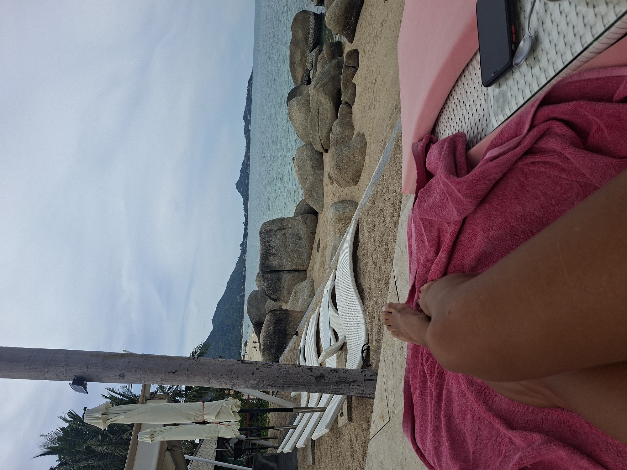The image depicts a perspective from a person lying on a pink towel on a lounger on a beach in Baan Hua Thanon, Thailand. The beach is sandy with large rocks scattered along the shoreline and in the water. In the background, a calm sea is visible with a distant island with hills and structures. The sky is overcast. To the left, there are several closed white umbrellas and a palm tree with a solar panel attached to its trunk. White lounge chairs are arranged on the sand. In the foreground, the legs and feet of the person are visible, resting on a pink towel. A pink lounger is partially visible on the right, with a smartphone and a spoon on a glass-topped table. The overall scene suggests a relaxed atmosphere on a cloudy day at the beach.