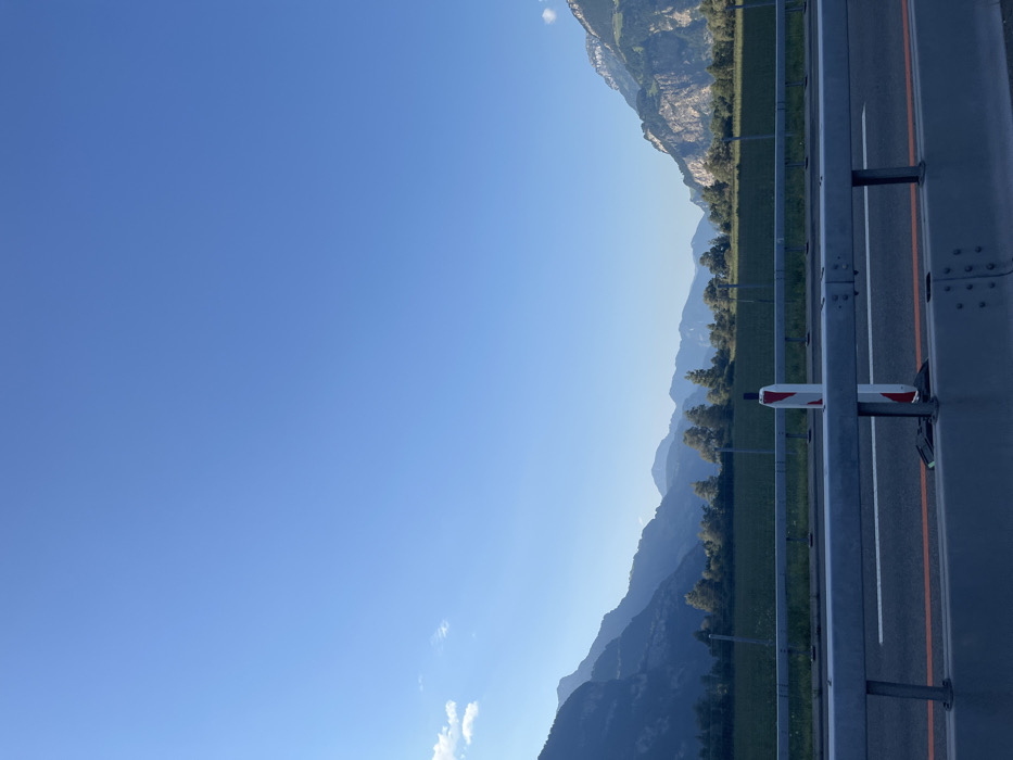 The scene depicts a static landscape near Seedorf (UR), Switzerland, viewed from a roadside vantage point. In the foreground, a two-tiered gray metal guardrail extends horizontally across the lower portion of the frame. Below the guardrail, a segment of asphalt road with a single orange-red lane marking is visible. Situated directly behind the guardrail and slightly to the right of the frame's center is a vertical white and red striped marker post. Beyond this marker and a wire fence, a flat, green agricultural field is present, populated by scattered trees that form a linear arrangement before the mountainous backdrop. The mid-ground and background are dominated by multiple layers of mountains, characterized by varied geological formations and receding atmospheric haze, with a distinct, rugged rock face visible on the right side. The upper half of the image consists of a clear, light blue sky, featuring a small cluster of white cumulus clouds in the lower-left quadrant. No individuals or dynamic actions are depicted.