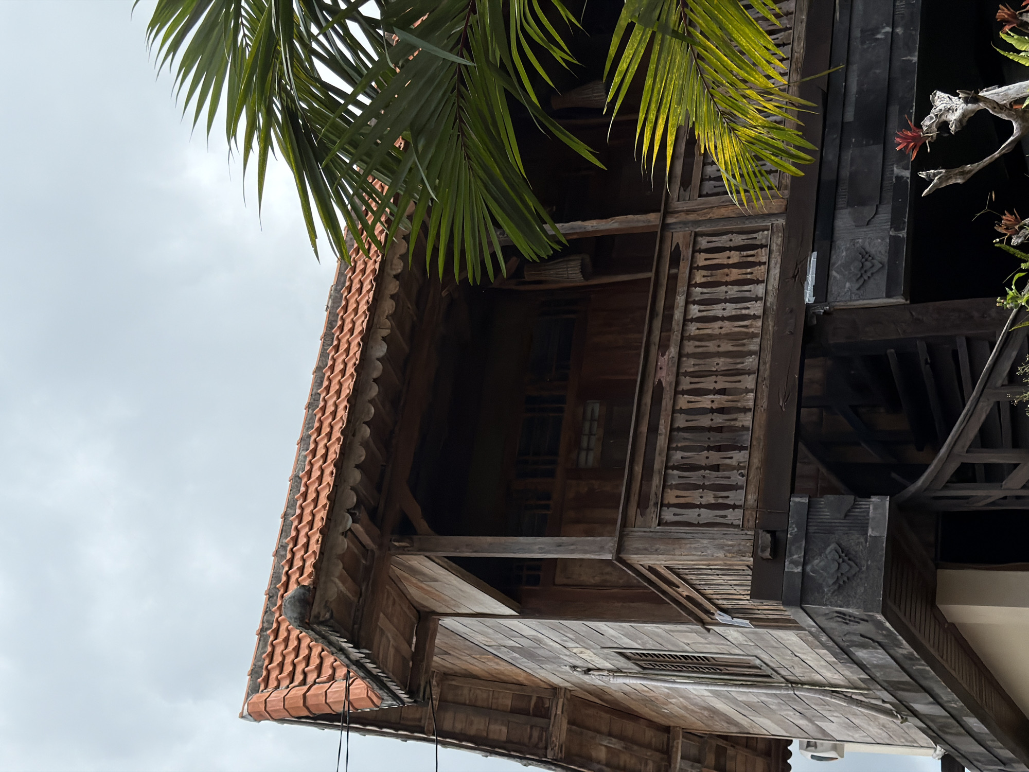 View of a three-story wooden building with a tiled roof in North Kuta, Indonesia, from a low angle. The building features intricate carvings and a dark, shadowed interior. Palm fronds partially obscure the structure, with an overcast sky in the background.