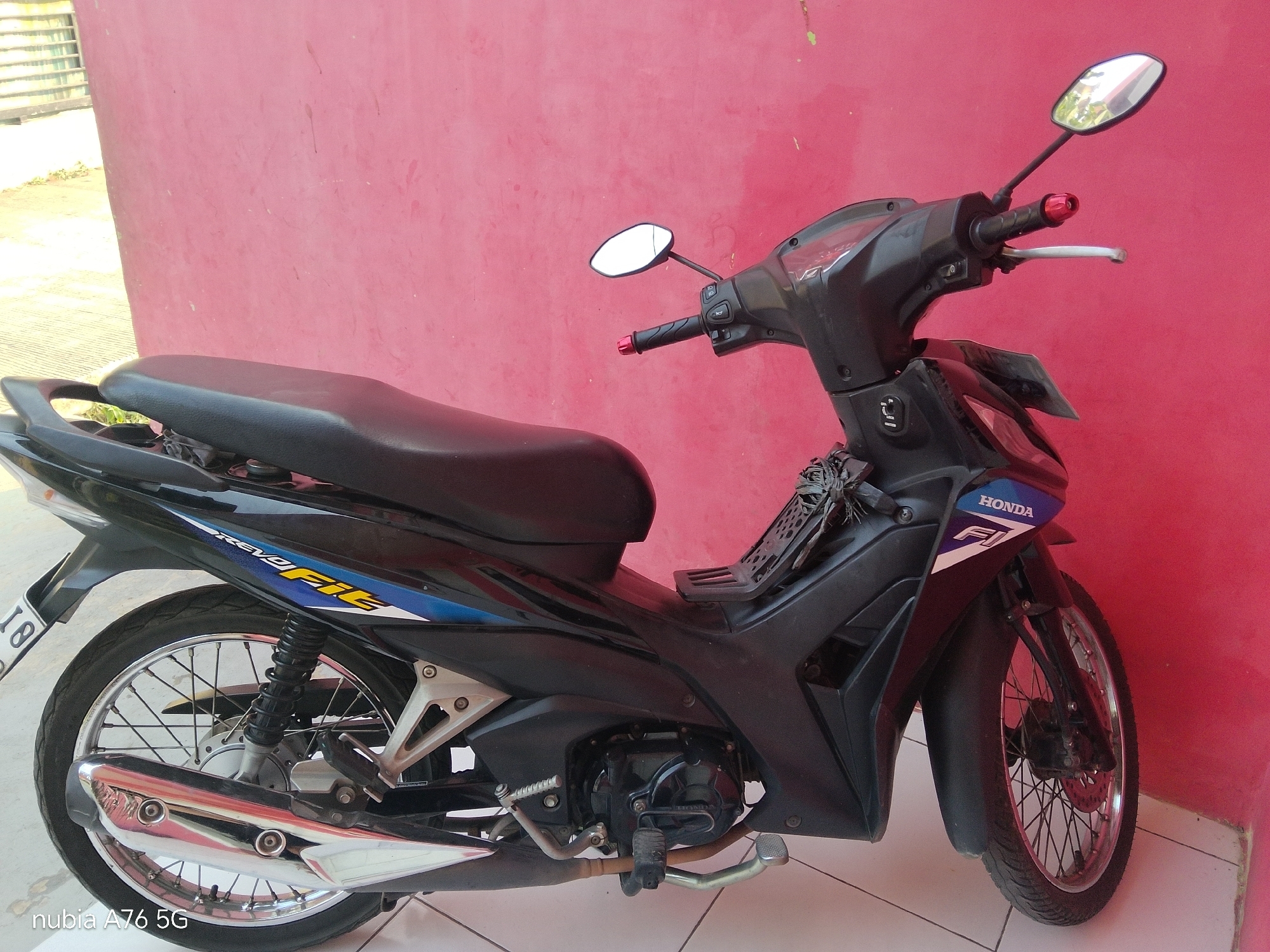 A black Honda motorcycle with "REVO FI" graphics is parked next to a vibrant pink wall. The motorcycle is positioned at an angle, with its front wheel slightly turned. It has a black seat, chrome exhaust pipe, and spoked wheels. The wall behind it is a solid, textured pink surface. The ground beneath the motorcycle appears to be tiled. The image has a watermark in the bottom left corner that reads "nubia A76 5G". The scene is outdoors, with soft lighting suggesting it might be daytime. The overall environment appears to be a residential or semi-urban area.