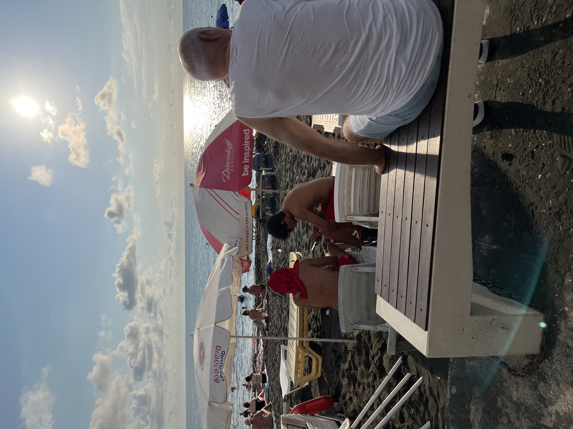 In Ureki, Georgia, on a dark sand beach, the late afternoon sun casts a bright glare on the Black Sea. The image captures a relaxed atmosphere with several people enjoying the beach. In the foreground, a man with gray hair and a white t-shirt sits on a wooden bench facing away from the camera, looking out towards the sea. To his left, another man, wearing a red headscarf and red shorts, sits on a white chair, looking down at his phone. Next to him, a young man in red shorts is also seated, looking at his phone.
Several beach umbrellas are visible, one white with red lettering and another red and white with "Davidoff be inspired" printed on it. Beach chairs and loungers are scattered across the sand, with some people sitting on towels near the water's edge in the middle ground. The sea is calm with gentle waves, and the sky is a clear blue with scattered white clouds. The overall scene depicts a typical day at the beach, with people engaging in leisure activities under the warm sun.