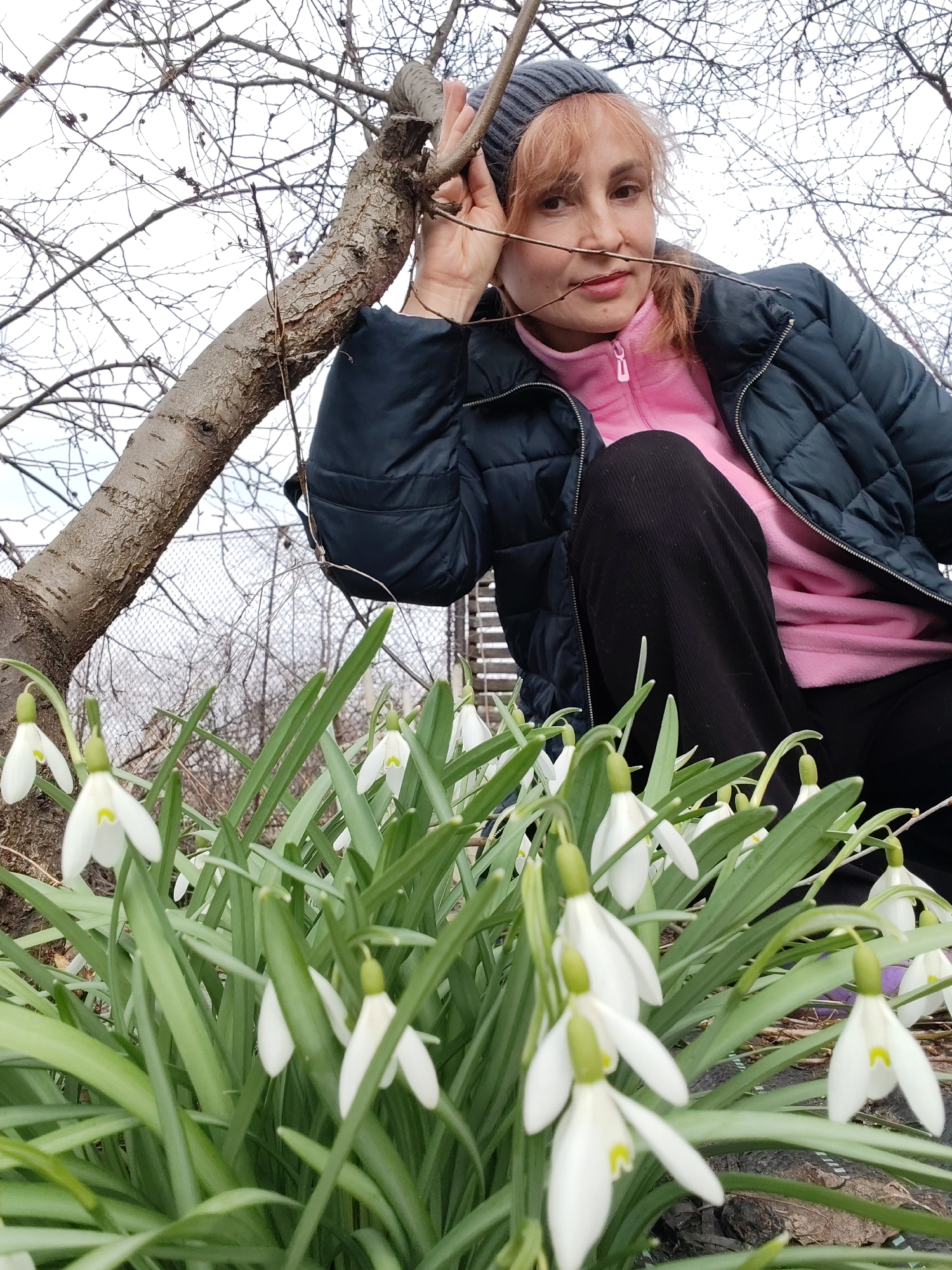 A medium shot shows a woman crouching next to a patch of snowdrop flowers outdoors in a garden setting. The woman is fair-skinned with light red hair and is wearing a grey knit hat, a pink fleece zip-up jacket, and a dark blue quilted jacket. She is also wearing black pants. Her left hand is near her face, lightly touching a small branch. The background is blurred but shows bare tree branches and a fence. The flowers in the foreground are white with green leaves. The location is Chaikovsky, Russia.