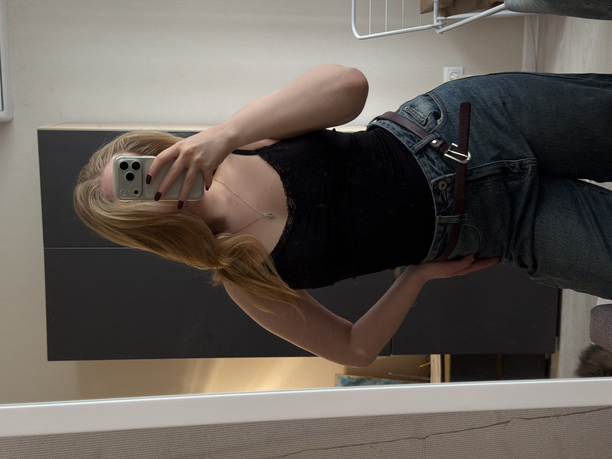 A young woman takes a mirror selfie. She wears dark wash jeans with a brown leather belt and a black tank top with lace trim. She is holding a smartphone up to her face, obscuring it. A silver necklace is visible. The background includes a gray cabinet, a white wall, and part of a clothing rack. The location is indicated as Засечное, Russia, but there are no specific details to confirm this.