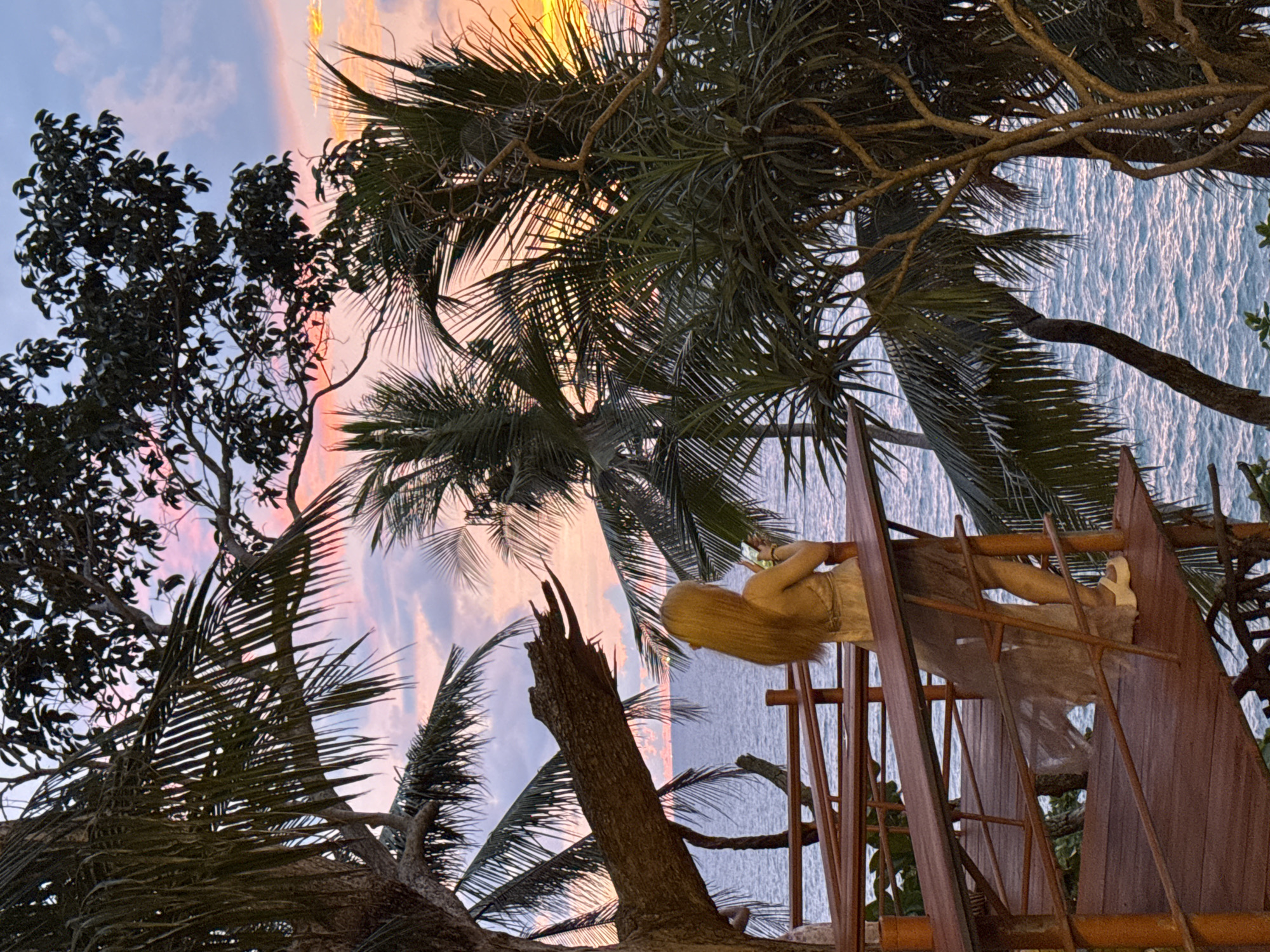 A woman with long blonde hair stands on a wooden platform overlooking the ocean at sunset in Karon, Thailand. She is wearing a light-colored dress and holding a phone in her right hand, appearing to take a picture or record a video. The platform is elevated and surrounded by lush palm trees, with their fronds framing the scene. The sky is painted with vibrant hues of pink, orange, and purple, reflecting on the calm sea. The overall atmosphere is serene and picturesque, capturing a moment of quiet enjoyment of the natural beauty.