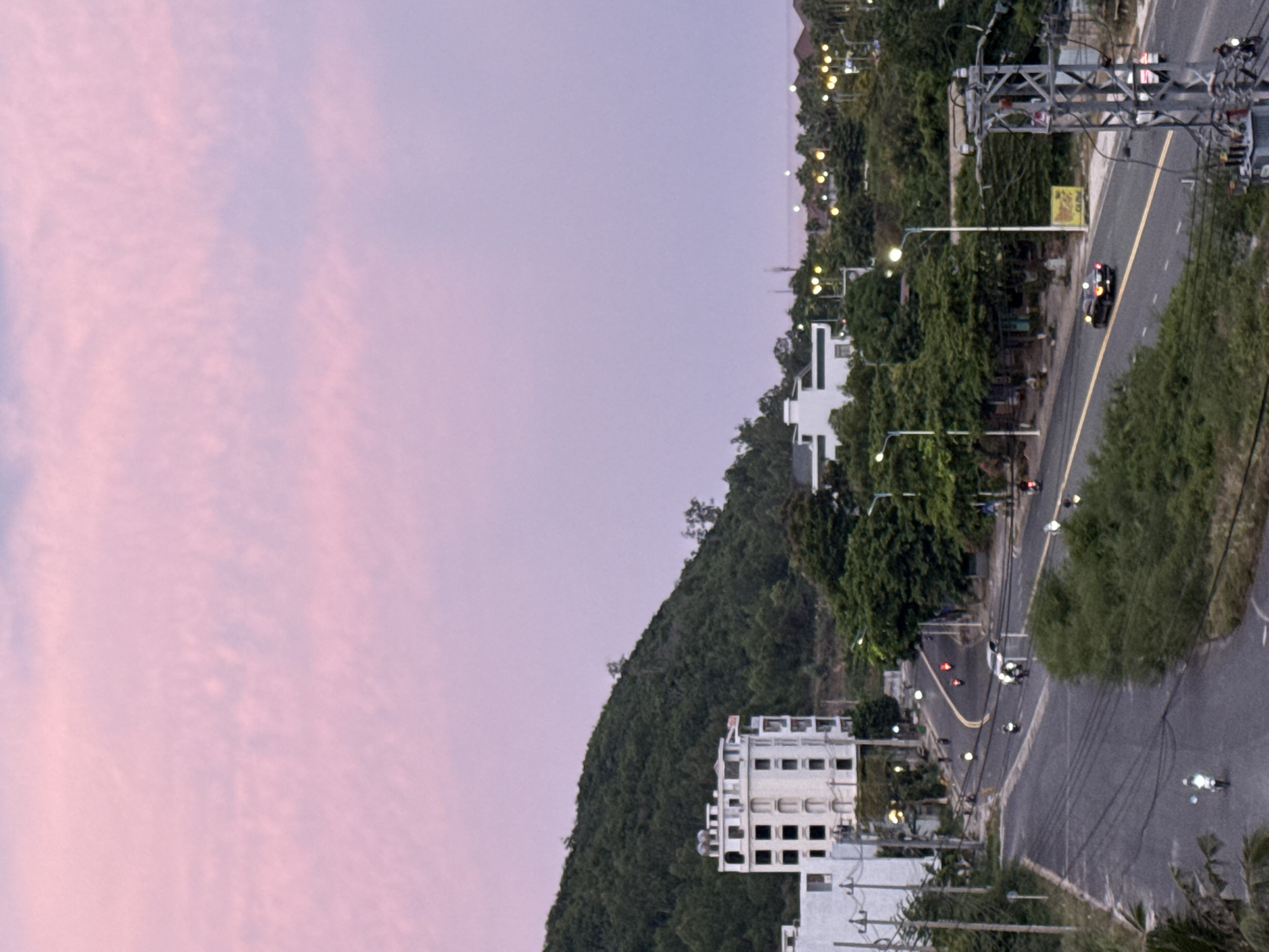This is a landscape photograph taken at dusk, with a soft pink and purple sky dominating the upper left portion of the frame. The image is oriented vertically. Below the sky, a lush green hillside slopes downwards. Several buildings are visible, including a multi-story white building with numerous windows situated on the hillside and another modern white building in the foreground. A road curves through the scene, with several motorcycles and a car visible on it. Streetlights and other lights from buildings illuminate the road and surrounding area, indicating it is twilight. The overall scene appears to be a coastal town or city, with the presence of a road and buildings suggesting an urban or semi-urban environment. The foliage indicates a tropical or subtropical climate. The time of day is clearly sunset or early evening, as evidenced by the colored sky and artificial lights. There is no discernible text in the image.