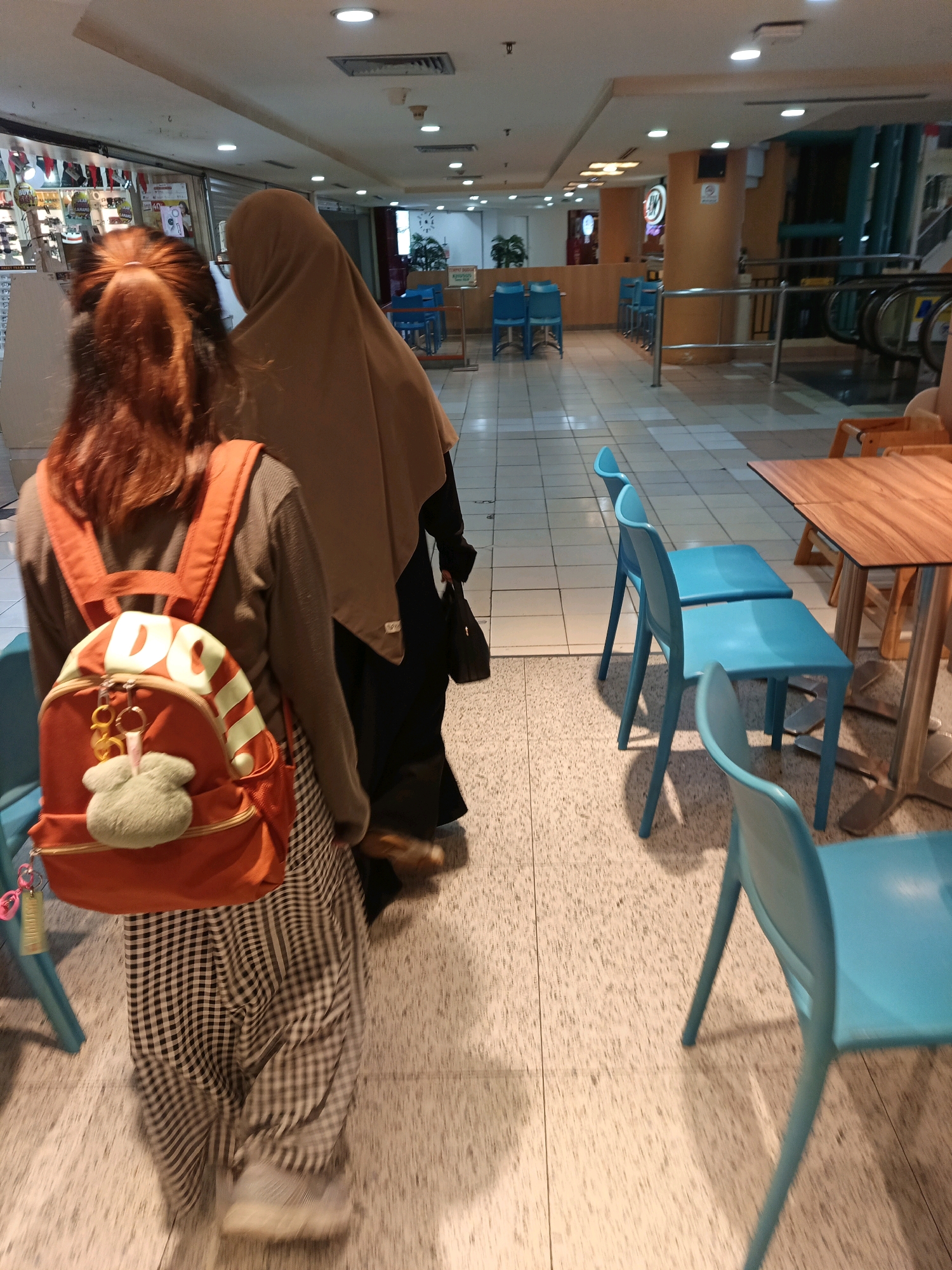 The image shows two individuals walking in an indoor public space, likely a mall or shopping center, in Central Jakarta, Indonesia. The person in the foreground is wearing a brown long-sleeved shirt, black and white checkered pants, and a bright orange backpack with a fluffy green charm. Their hair is red and tied up. The second person, partially obscured, is wearing a beige hijab and a black garment. They are walking away from the camera.
The environment features a tiled floor, a wooden table with metal legs, and several bright blue chairs. In the background, there are escalators, store fronts with displays of goods, and soft lighting from ceiling fixtures. The overall impression is of a modern, well-lit commercial area.