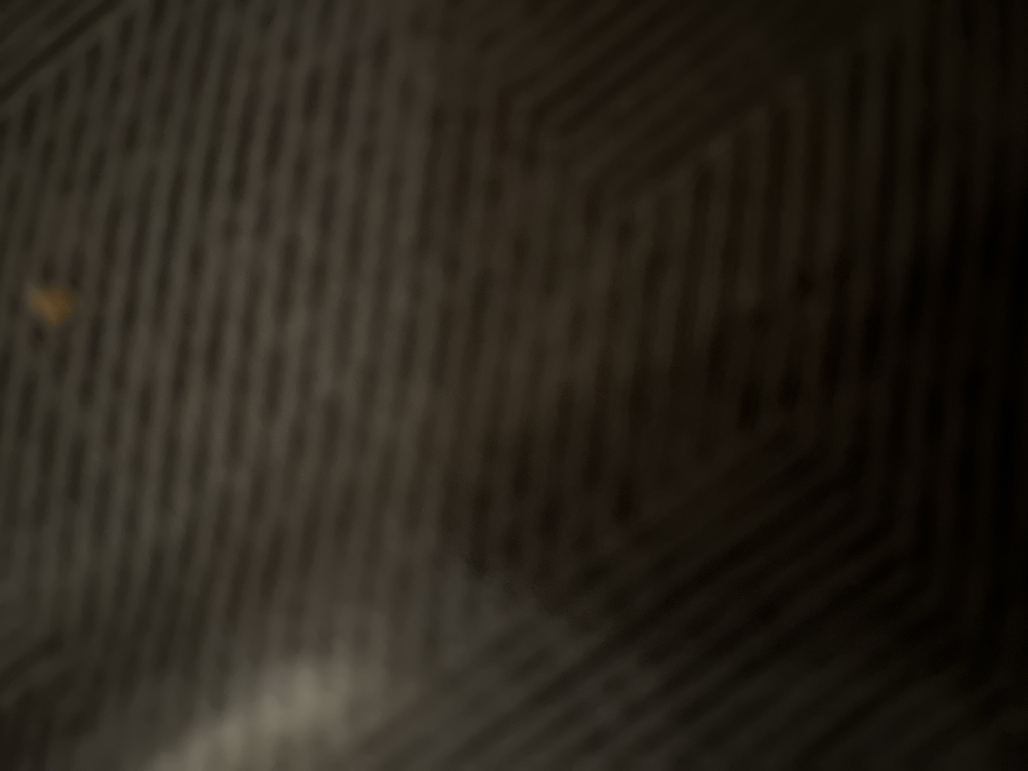 The image shows a close-up, blurry view of a dark, textured surface with a faint, light-colored smudge near the top center. The surface appears to have a repeating pattern of diagonal lines. The lighting is dim, creating shadows and obscuring details. The scene is not specific to Casablanca, Morocco, or any identifiable urban location. It is an abstract image lacking any recognizable landmarks or contextual elements.