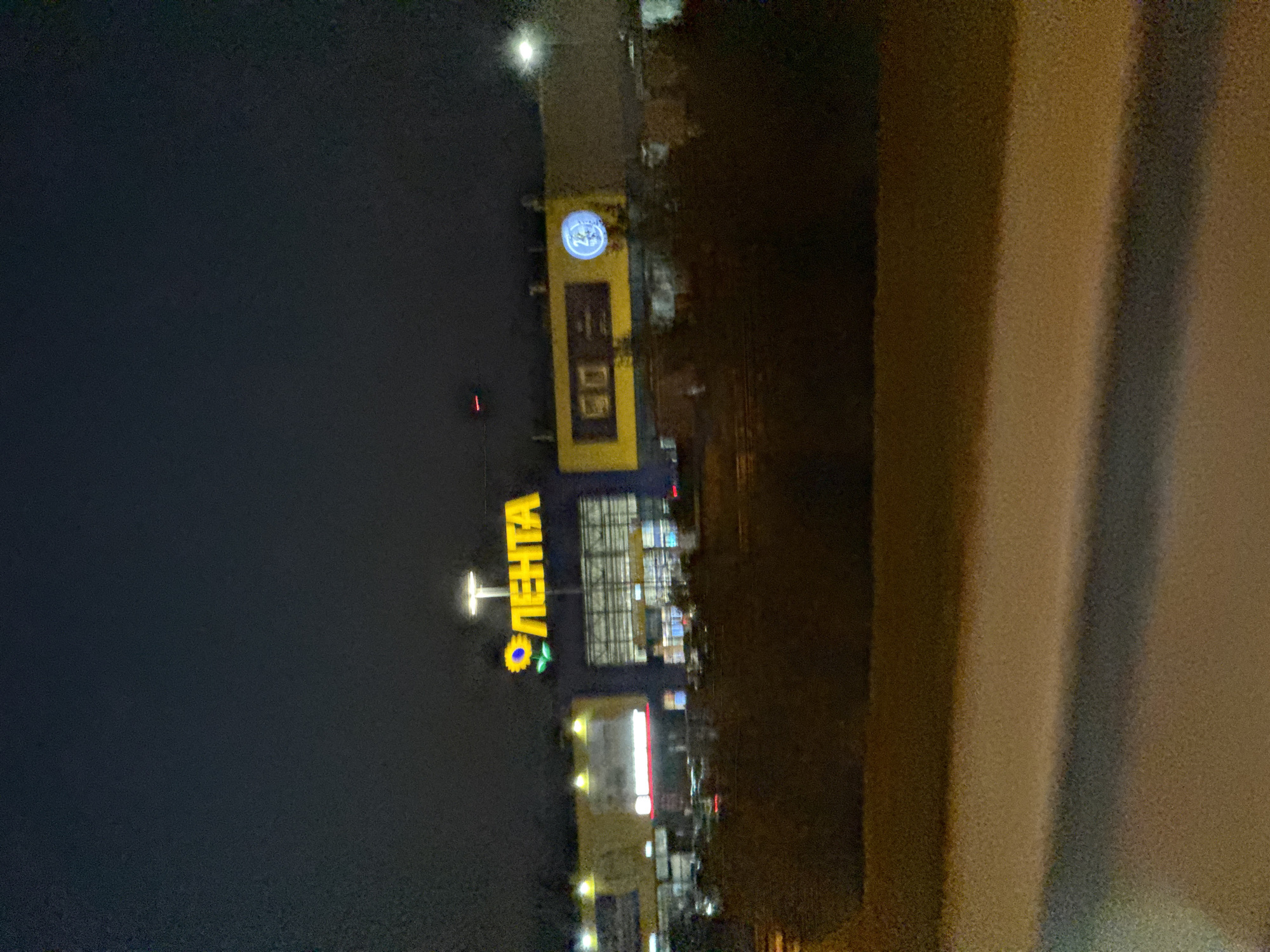 An exterior view of a Lenta supermarket at night, located at Staro-Petergofskoye Highway, 84, in Saint Petersburg, Russia. The building's facade is illuminated with bright yellow and blue signage, featuring the Lenta logo with a sunflower emblem and the word "ЛЕНТА" in large yellow letters. To the right, another section of the building is visible with yellow paneling and a circular blue illuminated logo. The surrounding area is dark, with some trees and foliage in the foreground. The image appears to be taken from a moving vehicle, as indicated by the slight blur and the presence of a blurred foreground element that resembles snow-covered ground or a road. The sky is dark and overcast, typical of nighttime in a city. There are no people or vehicles clearly visible in the scene.