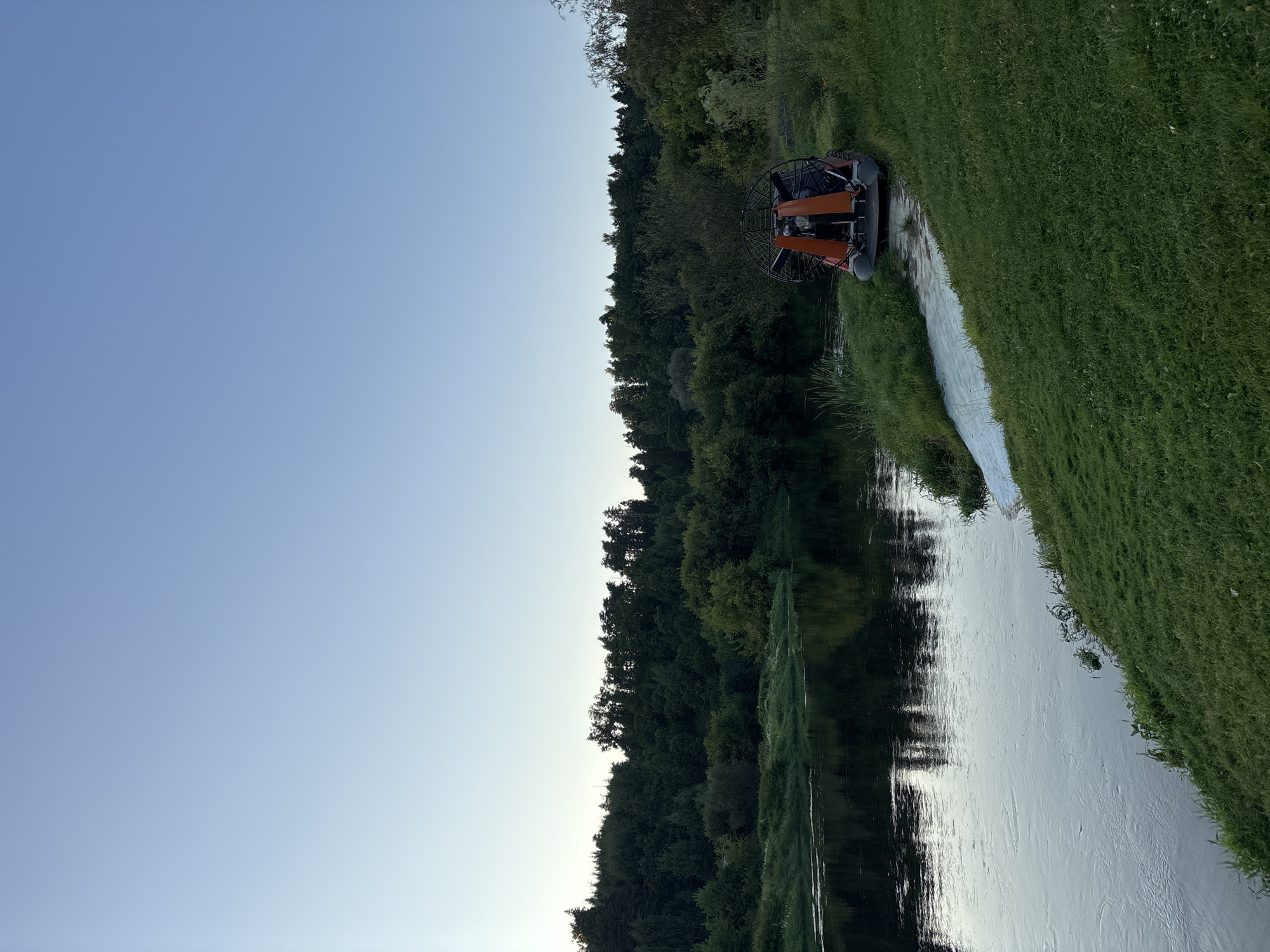 An airboat rests on the grassy bank of a wide, calm river with a dense forest lining the opposite bank and in the background. The sky above is a clear, pale blue, indicative of either early morning or late evening light. The airboat, predominantly orange and black, is positioned slightly to the right of the frame, facing the water. The river's surface reflects the surrounding greenery and the sky, creating a mirror-like effect. The grass on the bank is lush and green, with a white swath of material laid out near the airboat, possibly for launching or protection. The overall scene conveys a sense of quiet tranquility in a natural setting.