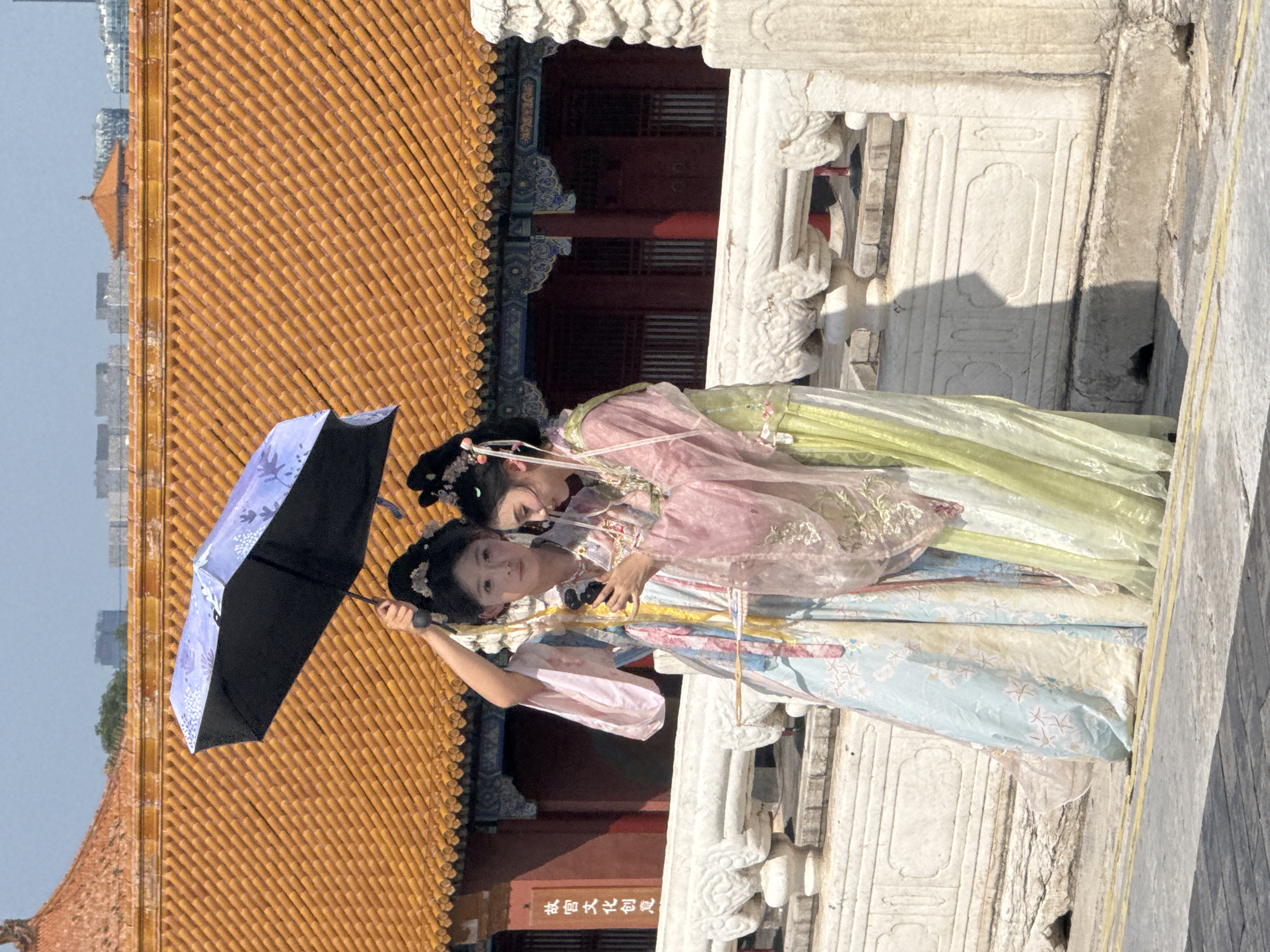 Two young women wearing traditional Chinese clothing and elaborate hairstyles stand on a stone walkway in front of a building with a yellow tiled roof. One woman, on the left, holds a purple umbrella over both of them. Both women are looking at a mobile phone held by the woman on the left. The woman on the right is wearing a pink and green layered robe, while the woman on the left wears a light blue and yellow robe. The architectural style of the building and the surrounding stone railings suggest the location is within the Forbidden City in Dongcheng District, Beijing, China. The sky is clear and blue.