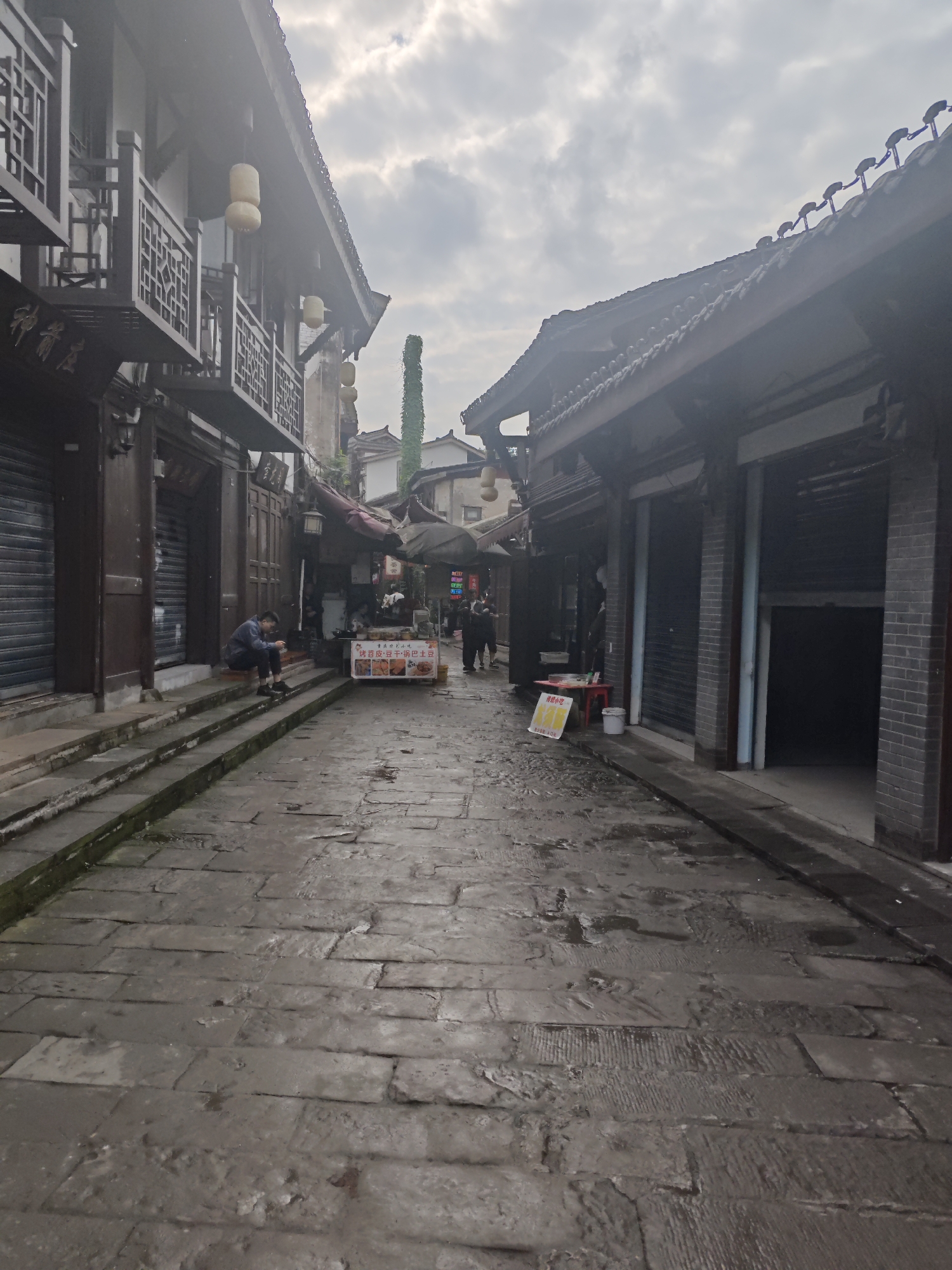 The image depicts a narrow, stone-paved street in Shapingba District, China. Buildings with traditional Chinese architectural features, including wooden balconies and tiled roofs, line both sides of the street. Many of the storefronts are closed with metal shutters. A man is sitting on the steps leading to one of the buildings. Further down the street, a food stall is set up, with a sign displaying food items in Chinese characters. Several people are visible in the background near the food stall. The sky is overcast, suggesting a cloudy day.