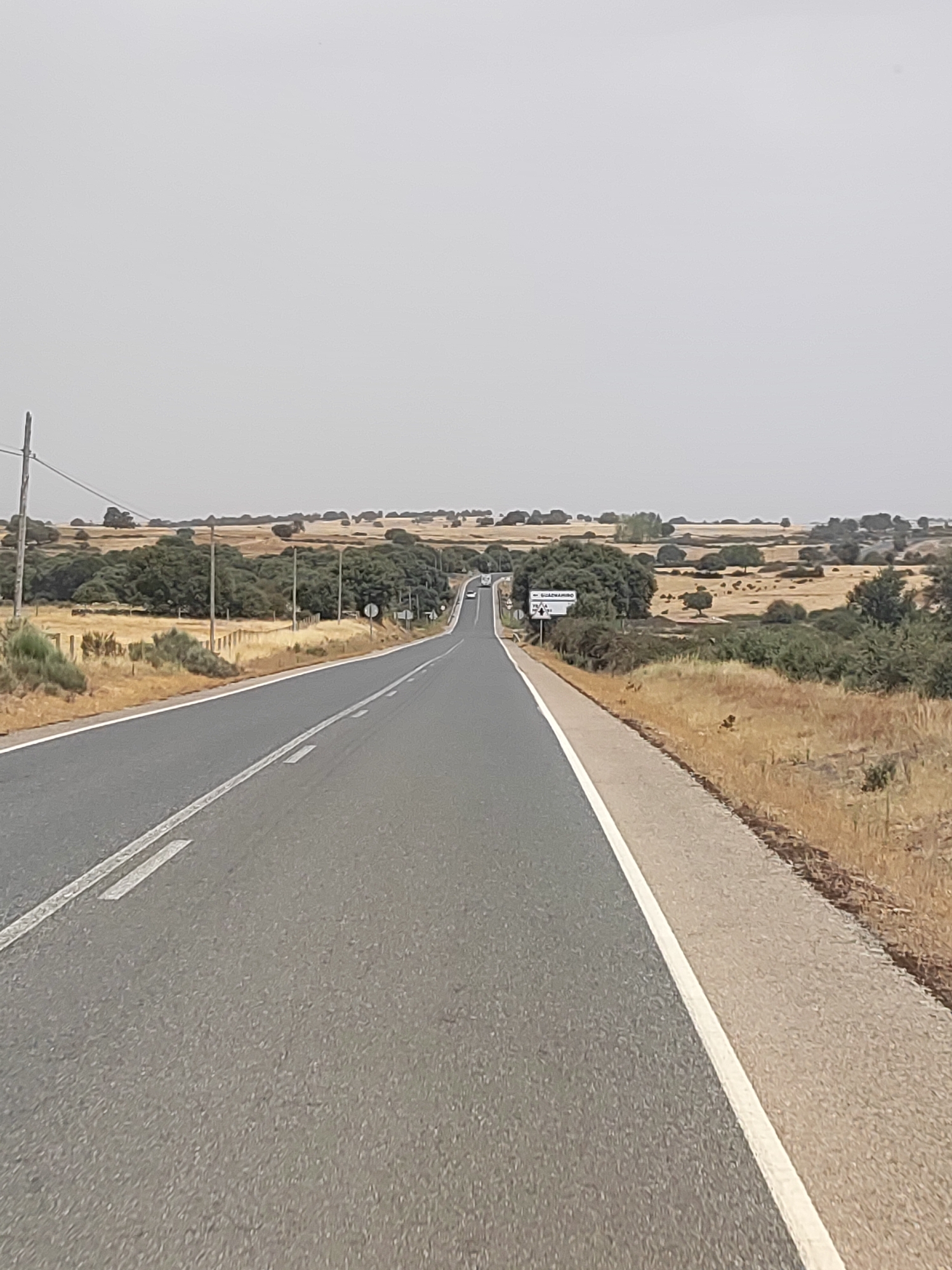 The image depicts a straight asphalt road receding into the distance, flanked by dry, golden fields and scattered trees. The sky is hazy and overcast, suggesting a warm, dry climate. In the mid-ground, a white sign with text in Spanish is visible on the right side of the road. The sign reads "- GUADRAMIRO -" followed by an arrow pointing left and the number "10". This indicates that Guadramiro, a municipality in the province of Salamanca, Spain, is located 10 kilometers in that direction. There are no people visible in the image. The overall scene conveys a sense of a rural, somewhat arid landscape on a highway leading towards a town.