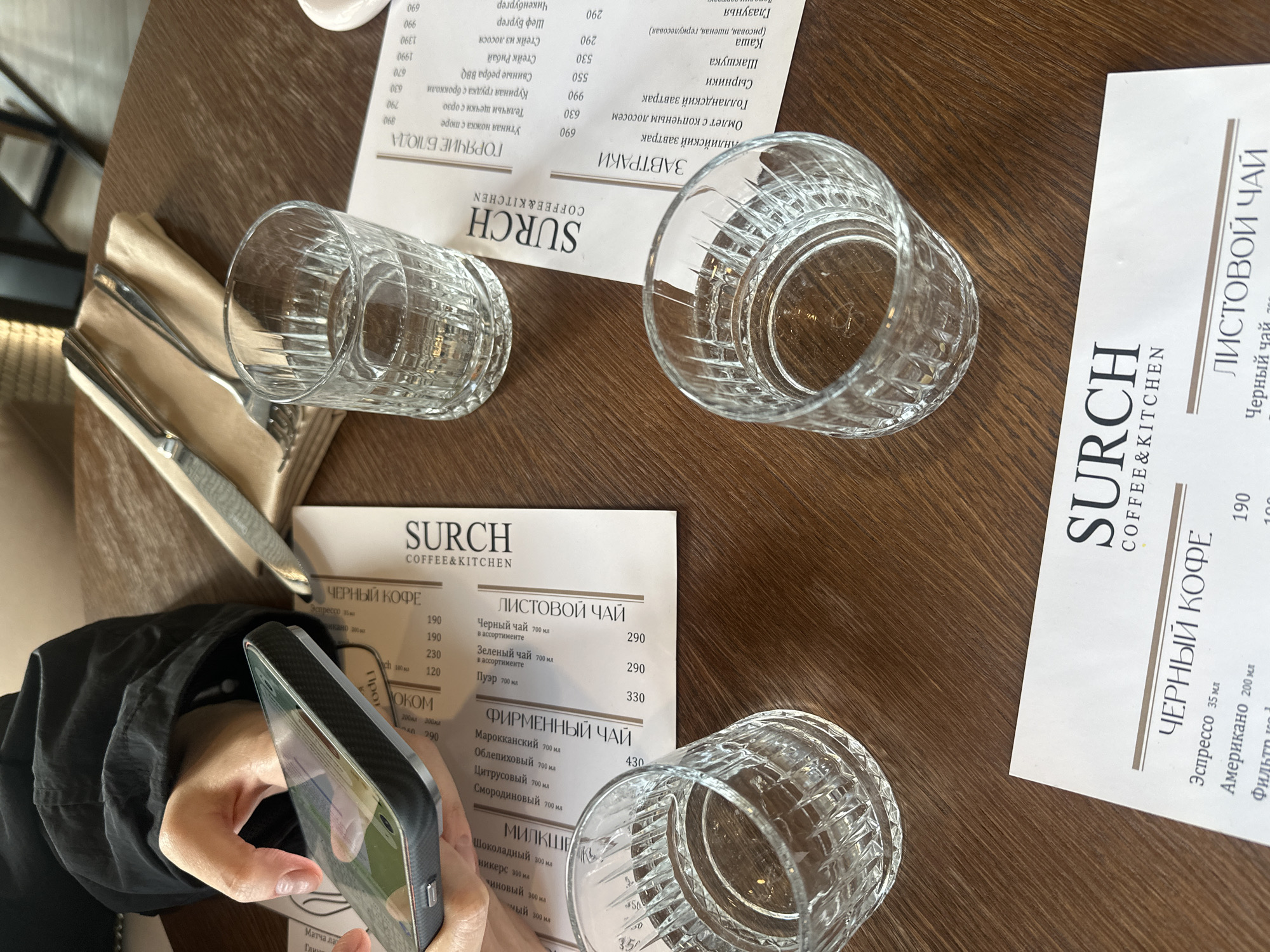 The image depicts a cafe or restaurant setting in Усады, Russia, viewed from an overhead perspective. A dark-grained wooden table occupies the majority of the frame. In the upper left, a person's left hand, clad in a dark, long-sleeved garment, holds a dark-cased smartphone horizontally. The phone screen displays reflections of the surrounding environment.
Three white menus, each bearing the logo "SURCH COFFEE & KITCHEN" at the top, are arranged on the table. They list various food and drink items in Russian, categorized under "ЧЕРНЫЙ КОФЕ" (Black Coffee), "ЛИСТОВОЙ ЧАЙ" (Loose Leaf Tea), "ФИРМЕННЫЙ ЧАЙ" (Signature Tea), "ЗАВТРАКИ" (Breakfasts), and "ГОРЯЧИЕ БЛЮДА" (Hot Dishes), with corresponding prices.
Three clear, empty glasses with a prominent vertical fluted design are distributed across the table. One glass in the upper center is partially filled with water, another in the lower left is also partially filled, and a third in the lower center appears empty. In the upper central area, a metal knife and fork rest on a folded beige napkin. The background is blurred, showing indistinct elements of the establishment's interior.