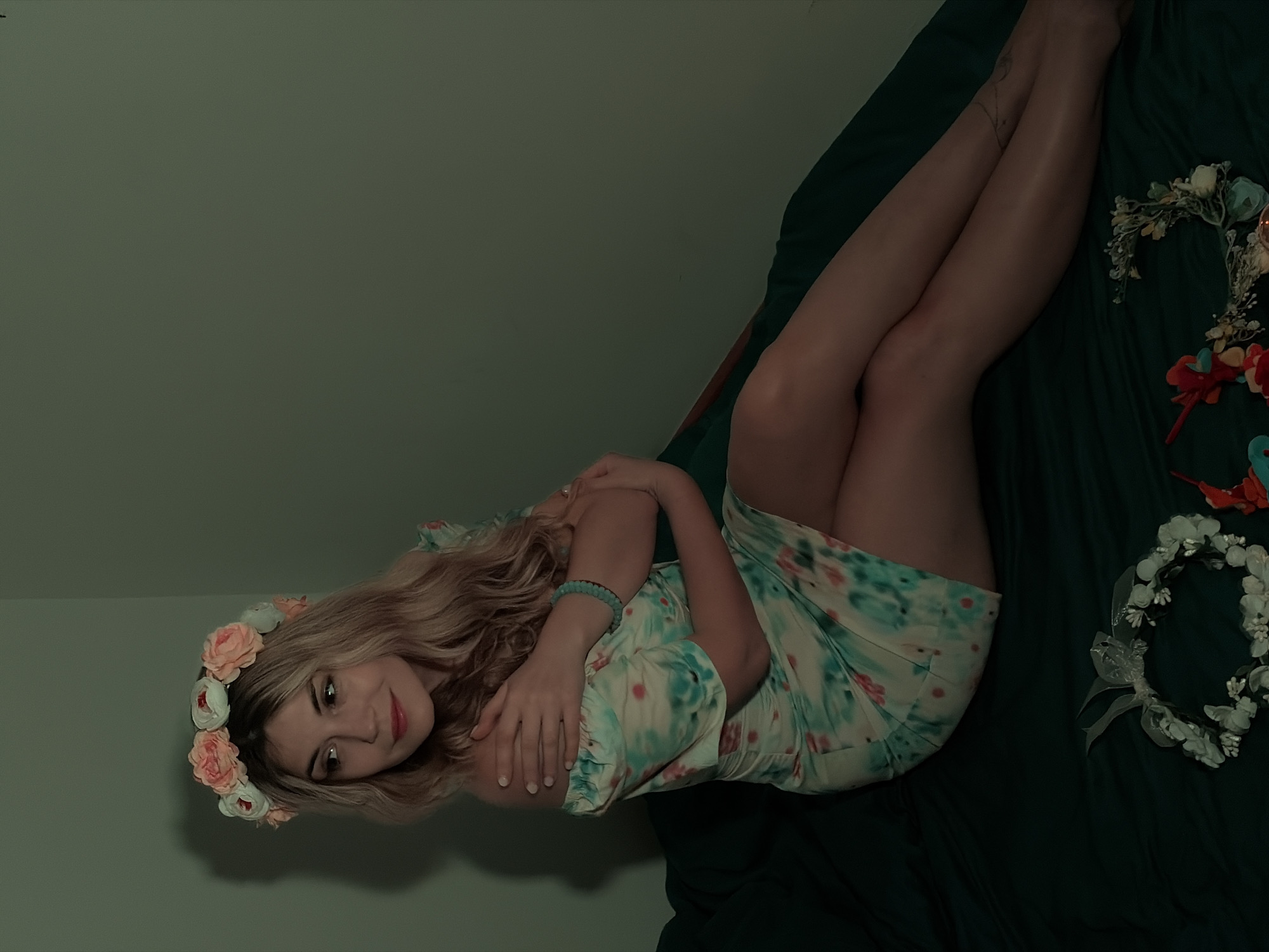 A young woman is lying on a dark green bed, wearing a floral dress and a flower crown. There are several flower crowns arranged on the bed around her. The background wall is a drab green color. The location is not identifiable as Budva, Montenegro based on the image content.
