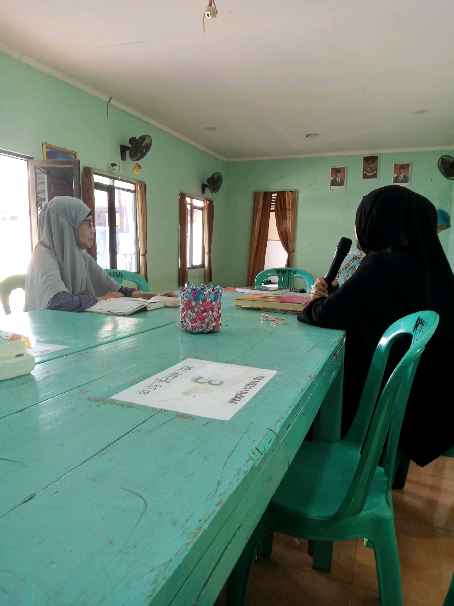 The image depicts an indoor scene, likely a classroom or meeting room, with two individuals present. The room has light green walls and a light-colored ceiling. The floor appears to be tiled with a light brown or beige color. Two windows with light brown curtains are visible.
On the left, a person wearing a light gray hijab and glasses is seated at a light teal wooden table, facing towards the right. They are wearing a long-sleeved garment. A book is open in front of them, and they appear to be reading or studying. Next to the book, a woven cylindrical object, possibly a pen holder or decorative item, is placed on the table.
On the right, another person is seated at the same table, facing left and away from the camera. They are wearing a black hijab and black clothing. This individual is holding a black microphone in their right hand and appears to be speaking.
The table is made of distressed light teal wood, showing signs of wear and tear. On the table, a piece of paper with some text and the number "3" is visible in the foreground.
Several items are mounted on the walls. On the left wall, a black oscillating fan is present. On the right wall, three framed pictures are displayed at different heights. The central picture appears to be a coat of arms, possibly the Garuda Pancasila, the national emblem of Indonesia. The other two pictures seem to be portraits of men in formal attire. Another black oscillating fan is mounted on the right wall.
Two green plastic chairs are visible, one partially obscured by the person in black on the right, and another on the far right edge of the frame. The overall impression is a simple, functional indoor space. The presence of the Garuda Pancasila suggests a connection to an Indonesian government or educational institution. The mention of "Cirebon, Indonesia" in the prompt suggests the location, but there are no explicit visual cues within the image itself that definitively confirm this specific city. However, the imagery aligns with typical Indonesian government or community meeting settings.