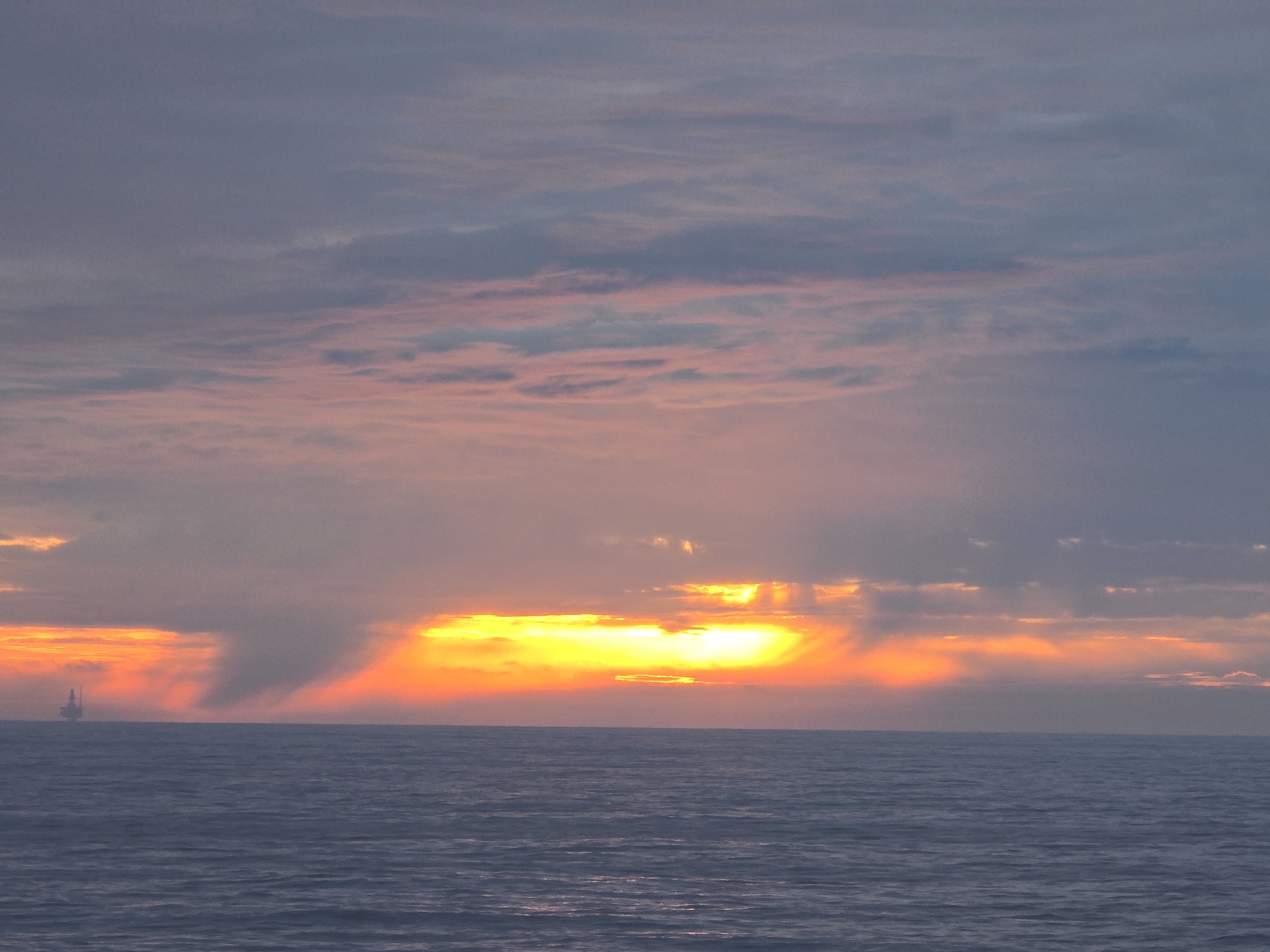 A seascape photograph captures a dramatic sunset over the ocean. The sky is filled with layers of clouds, transitioning from a soft pink and lavender hue in the upper portion to a bright orange and yellow near the horizon. The setting sun is partially obscured by the clouds, creating a radiant glow that illuminates the scene. A faint silhouette of what appears to be an oil rig is visible in the distance on the left side of the image. The ocean below is dark and wavy, reflecting the light from the sky.
Location: Unknown, Unknown.