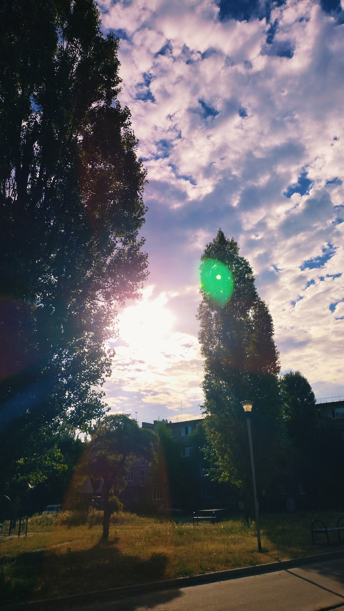 A bright, sunlit sky with scattered clouds is visible above a cityscape. Two tall trees, one on the left and one on the right, frame the sky. The sun is positioned between them, emitting a strong lens flare that creates a green circular effect on the right tree and a light ray effect on the left. In the foreground, a paved area leads to a grassy park with a small tree, park benches, and a lamppost. Buildings are visible in the background. The image appears to be taken in Horishni Plavni, Ukraine, judging by the architectural style of the buildings and the overall setting. There are no people present in the image.