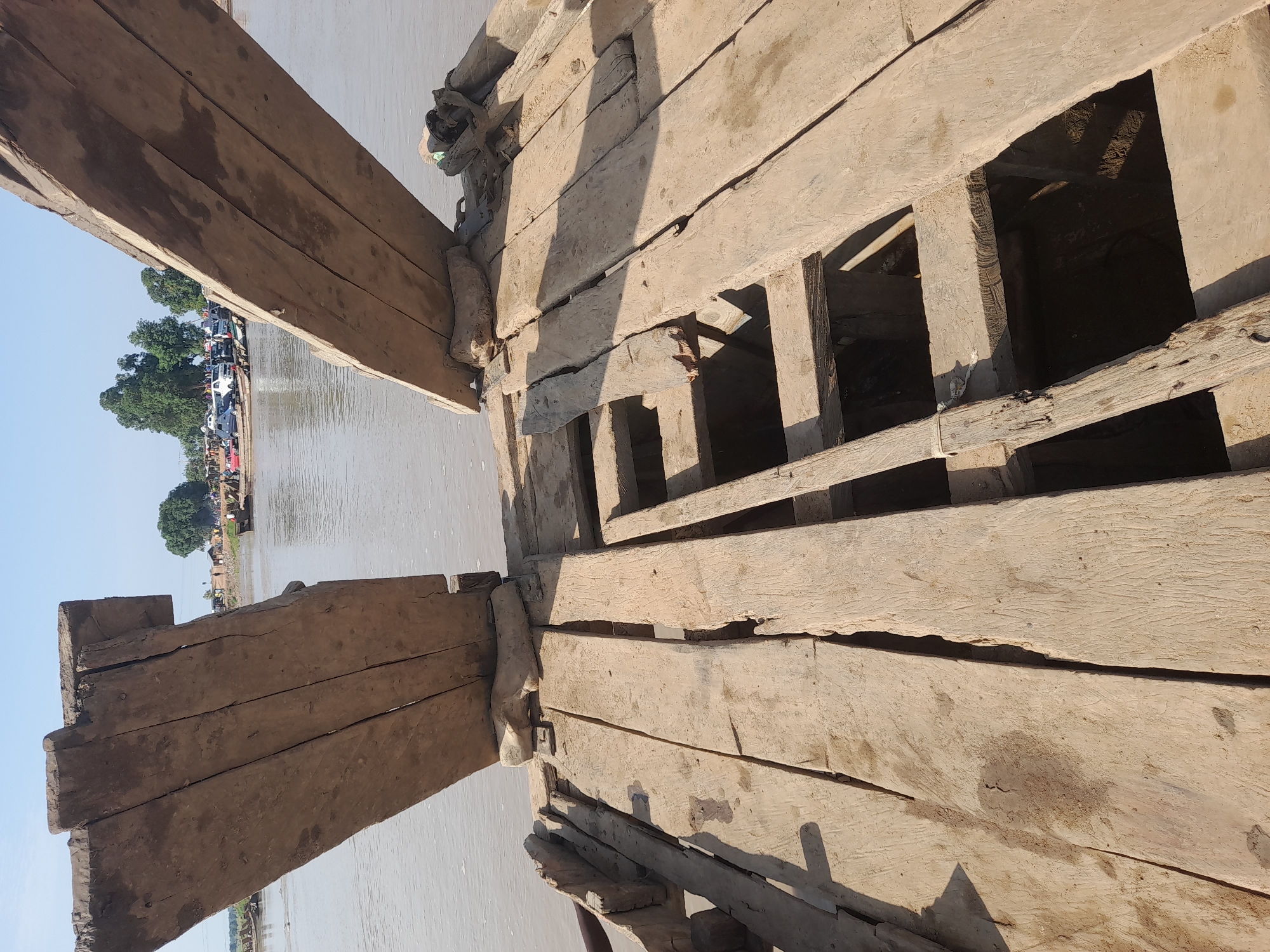 The image depicts a ferry crossing a river in Nemnei, Nigeria. The foreground is dominated by the weathered wooden deck of a ferry, revealing gaps and structural supports. Two large, angled wooden planks extend upwards from the ferry deck, forming a makeshift ramp or entryway. In the background, the murky river flows towards a far bank lined with trees and a small settlement. Several other ferries are visible on the water, some laden with vehicles, including cars and what appear to be trucks. Numerous people are gathered on the ferries and the riverbank, suggesting an active transportation hub. The overall scene conveys a sense of rudimentary but functional riverine transport.