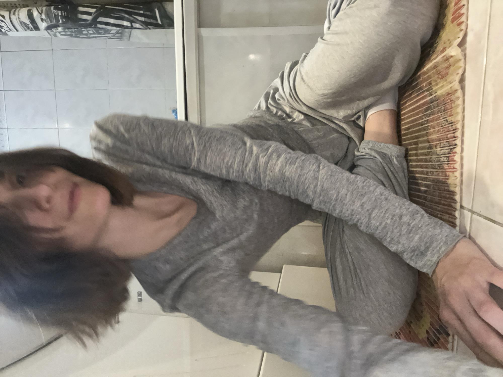 A woman with short brown hair is wearing gray sweatpants and a long-sleeved gray shirt. She appears to be seated or lying on a surface next to a wall. The setting seems to be indoors, possibly a bathroom or room with tiled walls. The image's perspective is slightly tilted. The location is not discernible from the visual information provided.
