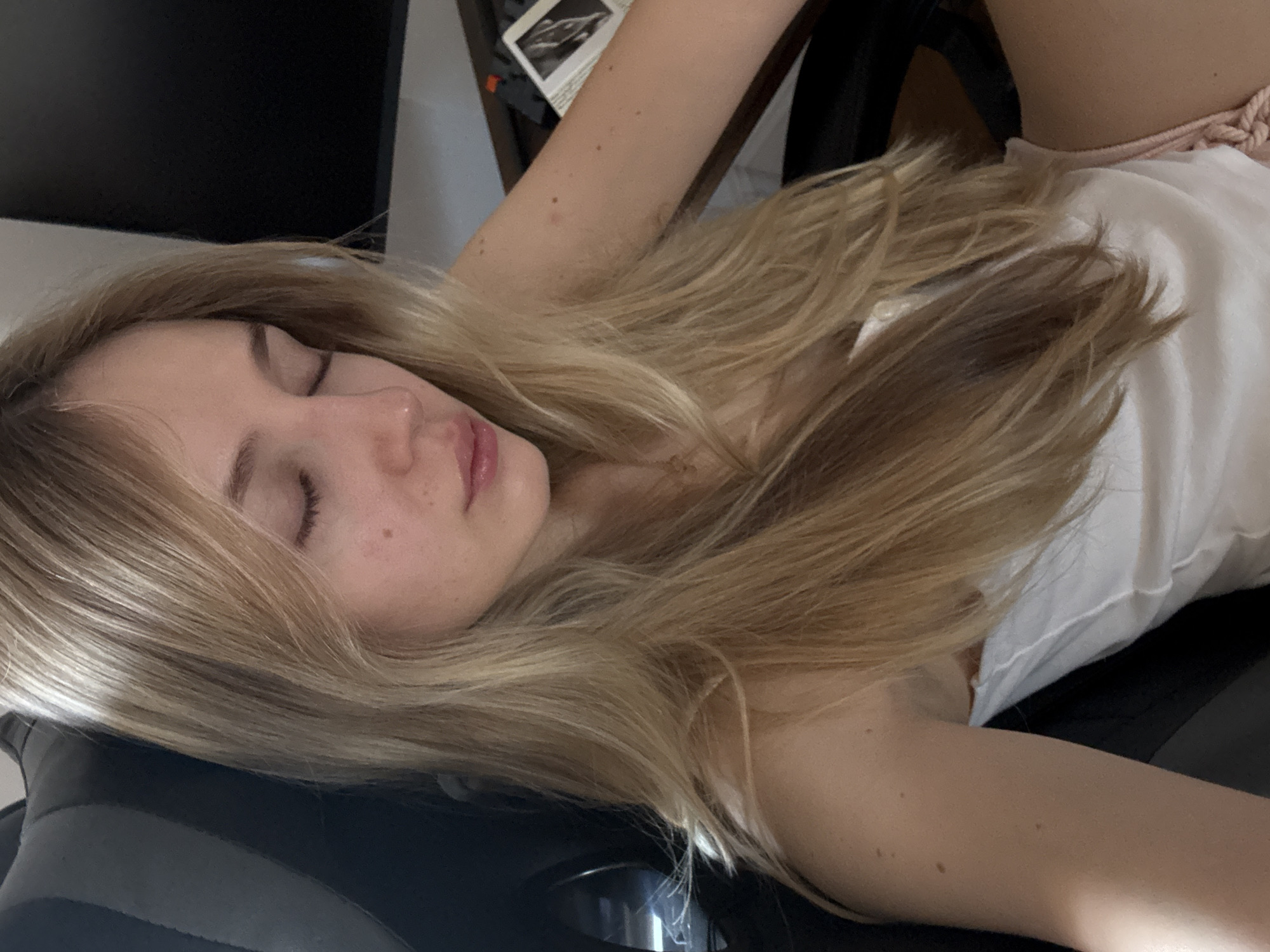 The image presents a close-up portrait of a young woman with long, light blonde hair, appearing to be asleep or deeply resting. Her eyes are closed, and her facial expression conveys a sense of serenity and peace. Her fair skin shows visible freckles or moles, particularly on her face and arms.
She is wearing a white top or tank top, with a braided, light pink or beige strap visible over her right shoulder. Her head is comfortably positioned on a dark, upholstered surface, which appears to be the headrest of an office or gaming chair.
The lighting in the scene is soft and ambient, indicating an indoor setting. In the blurred background, a dark, flat object, possibly a monitor or screen, is visible. Adjacent to it are some indistinct papers, one of which contains a very blurry, unidentifiable image. The overall activity depicted is one of quiet repose or sleep.
No specific time of day or outdoor weather conditions can be determined from the indoor environment. There is no visible, legible text within the image. The setting for this scene is Mexico City, Mexico.