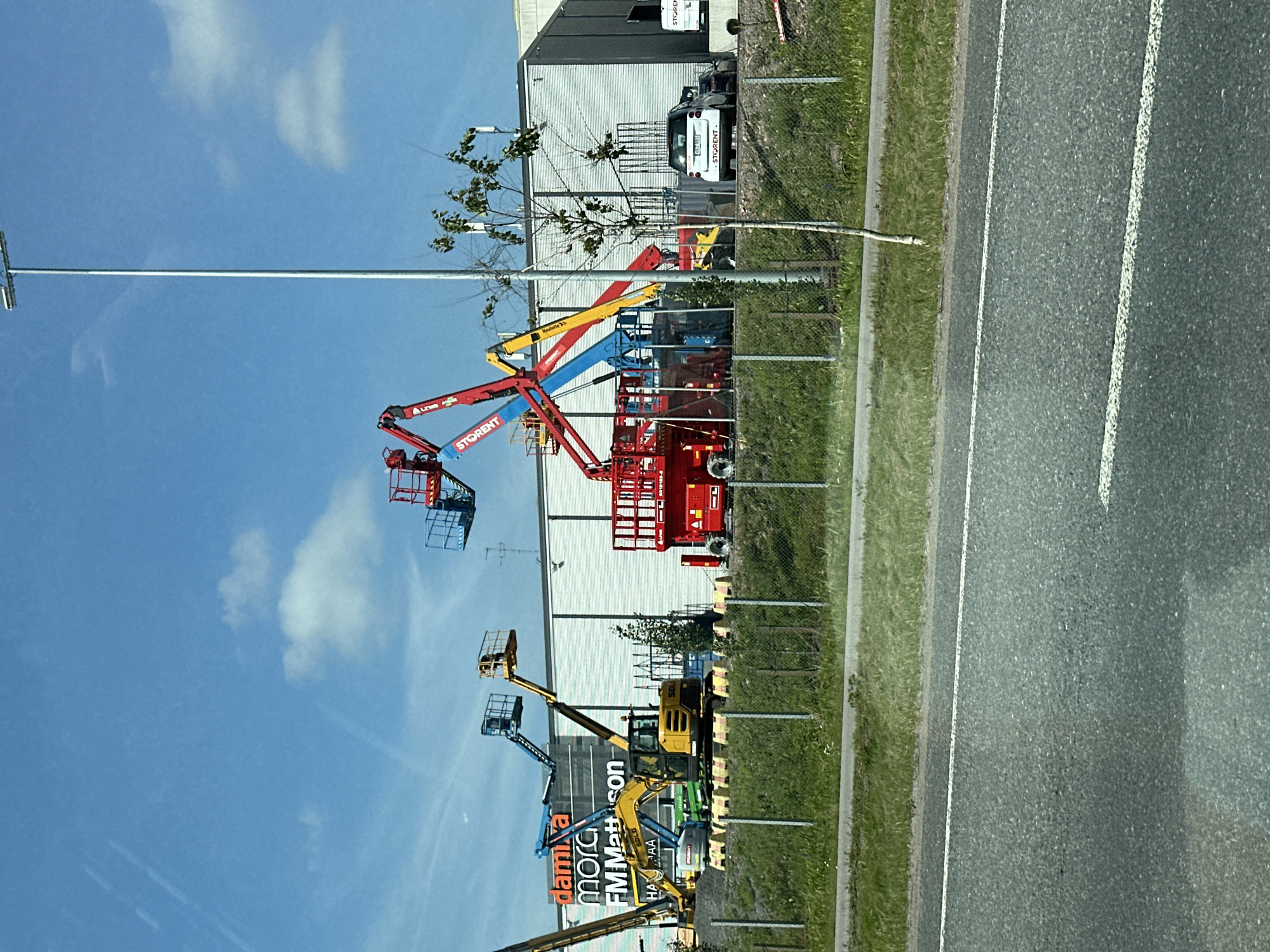 This is a medium shot, eye-level, outdoor, daytime photograph taken from a moving vehicle. The image depicts a row of construction vehicles and equipment lined up in front of a large, corrugated metal building under a blue sky with scattered white clouds. The building has a white facade and a dark roof. Several boom lifts, in various colors including red, yellow, and blue, are parked in front of the building. A yellow excavator is positioned to the left of the boom lifts. A white car with a license plate is visible to the right of the equipment, along with another dark car parked behind it. The scene is bordered by a metal fence and green grass in the foreground, with a paved road in the immediate foreground. The lighting suggests a sunny day.