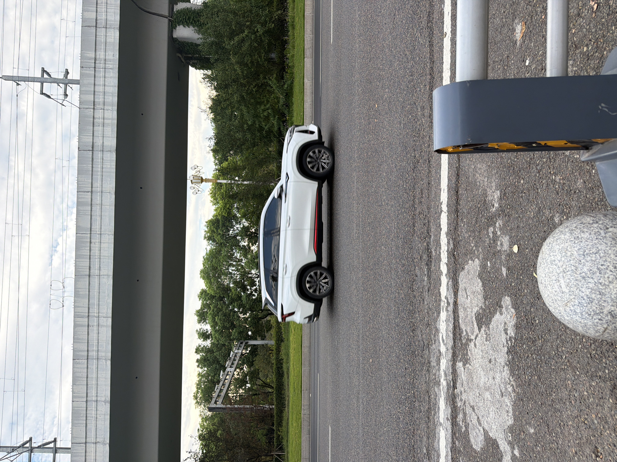 A white SUV with red trim drives on a paved road under a concrete overpass. The overpass has power lines running across its top. Trees and a street lamp are visible behind the road. In the foreground, a gray sphere and a dark gray barrier with yellow accents are visible on the side of the road. The sky is cloudy. The scene appears to be located in Jiangjin, China, based on context.