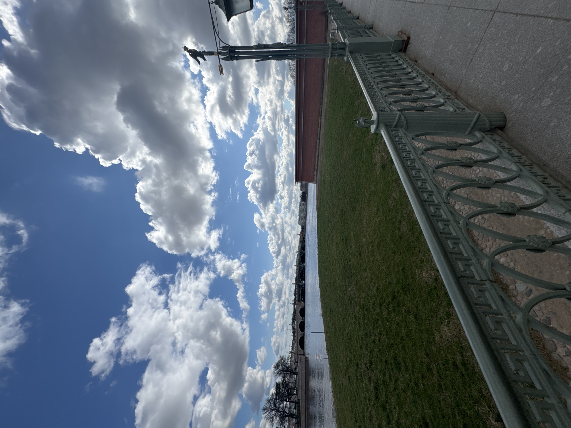 The image depicts a scene in Saint Petersburg, Russia. The perspective is a ground-level view, looking up at a sky filled with white clouds against a blue sky. A decorative, green-painted metal railing runs along the right side of the frame, next to a stone walkway and green grass. A red brick wall is partially visible behind the grass. In the distance, a bridge spans a body of water, and bare trees line the waterfront. A lamppost is also visible. The scene is set during the daytime.