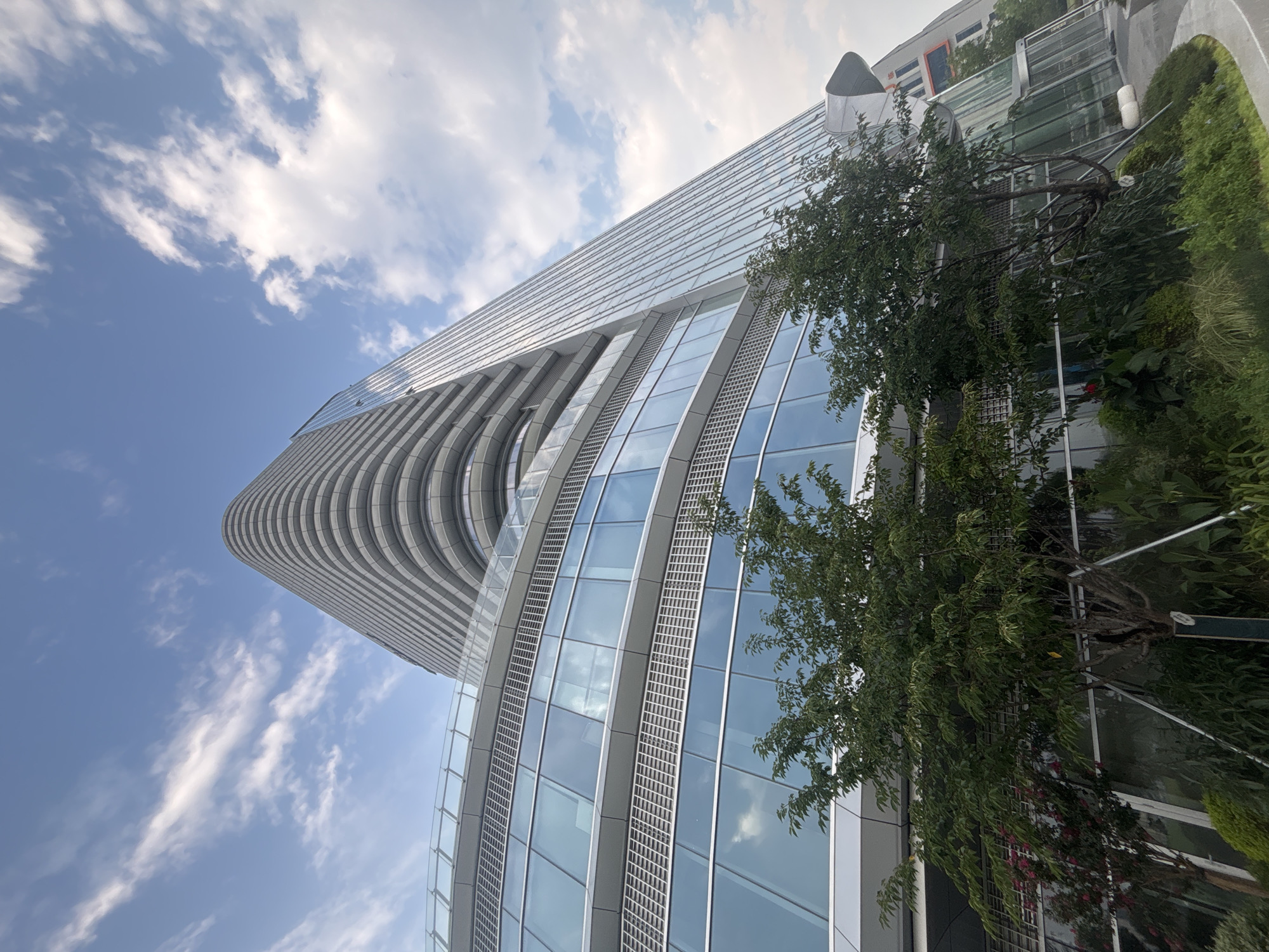 The photograph captures a modern, curved skyscraper in Suzhou Industrial Park, China. The building features a sleek, glass facade with horizontal bands of reflective panels and is angled upwards, creating a dramatic perspective against a partly cloudy blue sky. Lush green trees and landscaping are visible in the foreground, softening the imposing architecture. The scene is static, with no people or activity depicted. The lighting suggests it is daytime, possibly late afternoon given the slightly diffused sunlight.