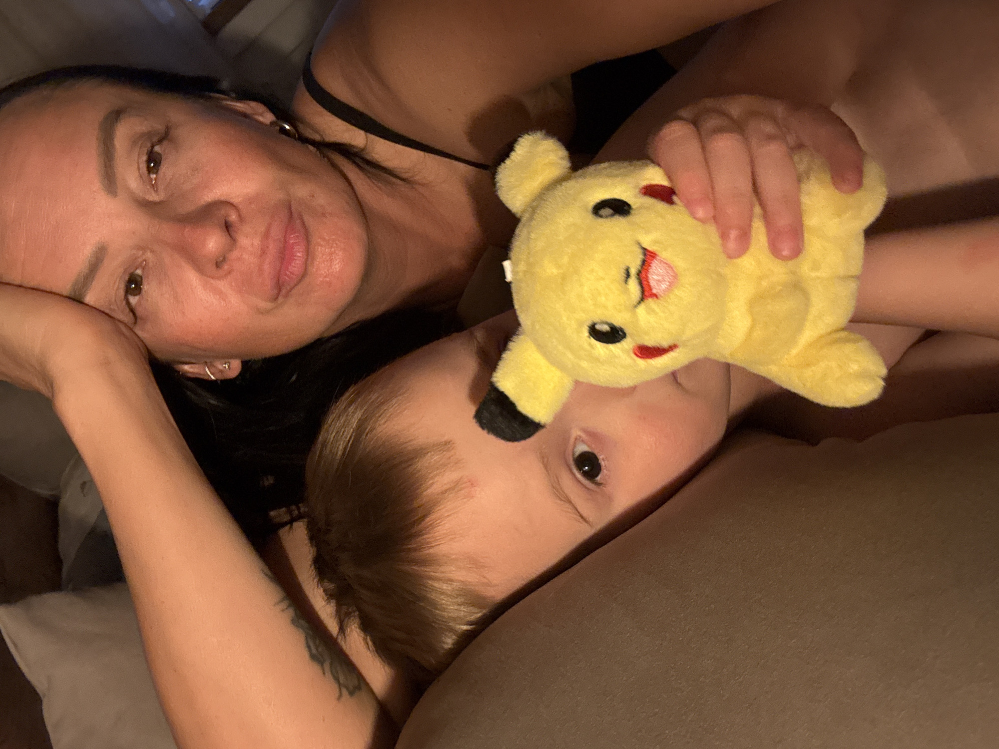 A woman and a child are lying down together, likely in a bed. The woman is at the top of the frame, resting her head on her left hand, looking at the camera with a neutral expression. She has dark hair and visible ear piercings. A black strap is visible, indicating she is wearing a tank top or similar garment. The child is positioned below her, partially obscured by the woman's arm and a stuffed Pikachu toy that the woman is holding. The child has light brown hair and is looking to the side. A tattoo is visible on the woman's left arm. The environment appears to be a dimly lit bedroom or living space. There is no clear indication that the location is the city of Chaikovsky, Russia.