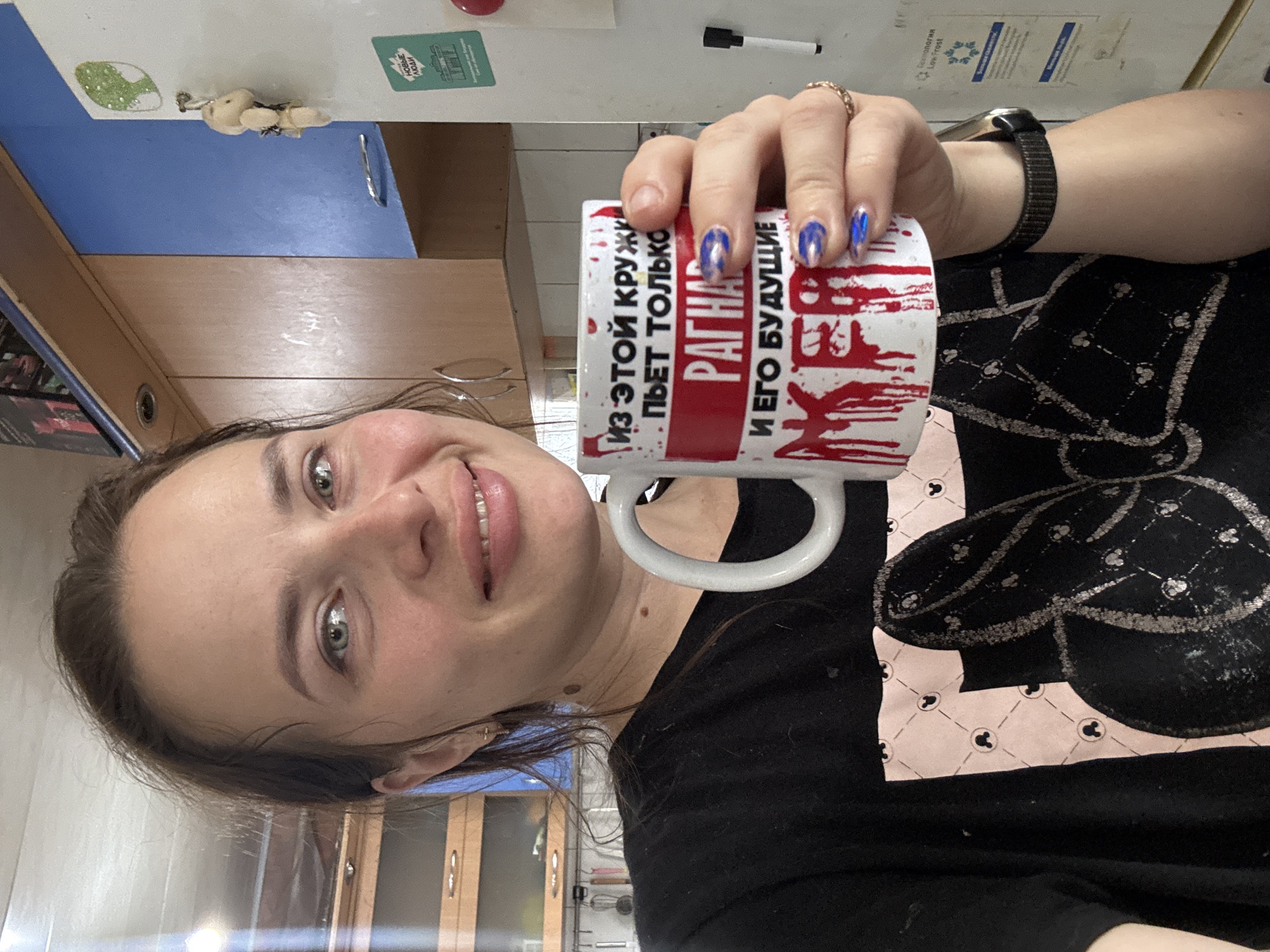 A woman with light brown hair and blue eyes is holding a white mug with red lettering and blood-like splatters. She is wearing a black t-shirt with a pink and black graphic of Minnie Mouse ears. The mug's text reads "Из этой кружки пьет только Рагнар и его будущие жер". The background shows a kitchen with wooden cabinets, a blue cabinet, and a white refrigerator with a sticker and a small stuffed toy hanging from it. The location is identified as Rusalevka, Russia.